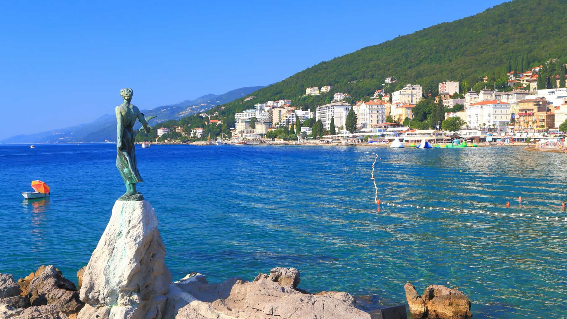 A beautiful coastal shot of Opatija, Croatia, with the iconic "Maiden with the Seagull" statue on a rock in the foreground, facing the clear Adriatic Sea and a bustling waterfront promenade.