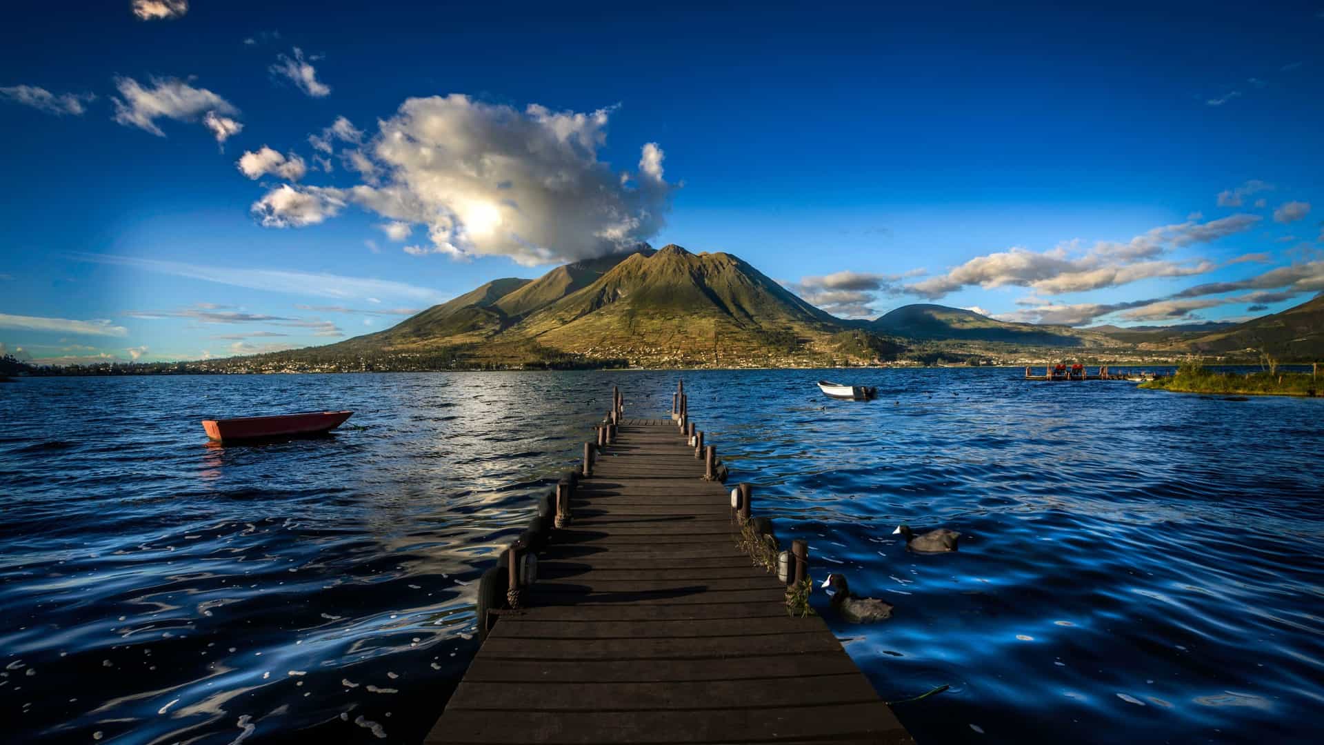 A long wooden pier extends into the deep blue waters of Lake Atitlán in Guatemala, with a majestic volcano in the background under a vibrant blue sky.