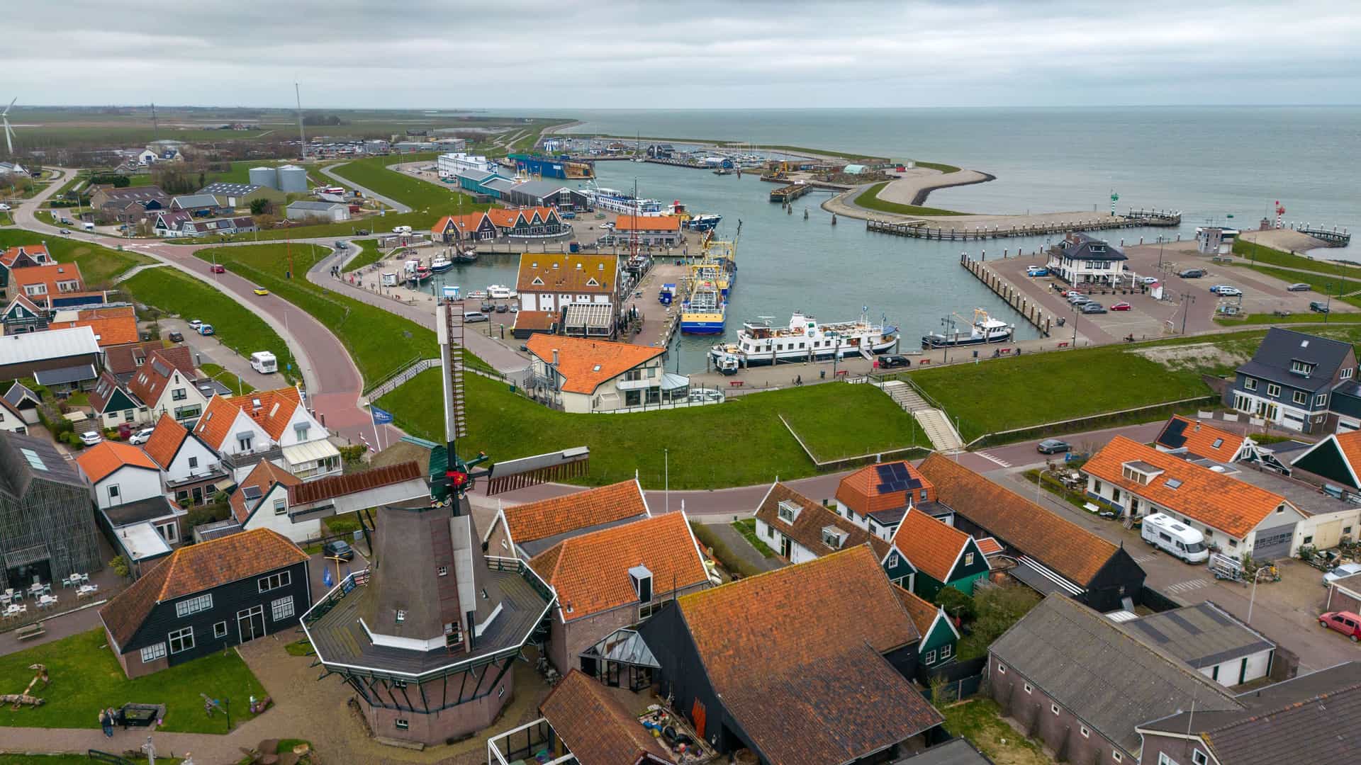 "An aerial shot of the serene fishing harbor in Oudeschild, the Netherlands, with boats docked and a windmill with a red roof in the foreground.  "