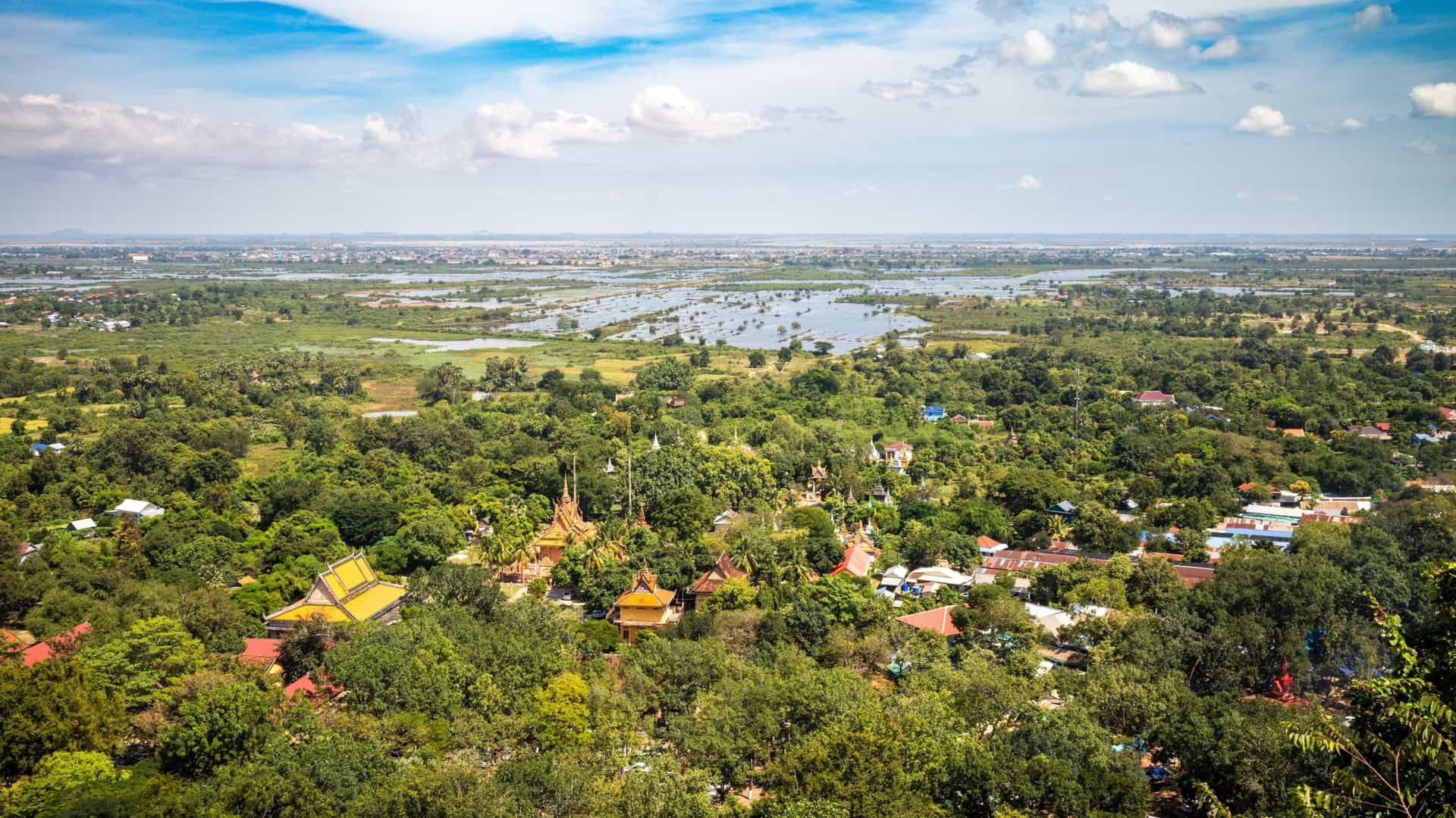A panoramic view from the top of Phnom Oudong (Oudong Temple) in Cambodia. The image captures the vast, flat landscape of the Cambodian plains and rice fields stretching out to the horizon under a pale, cloud-streaked sky. In the immediate foreground, the architecture of the Buddhist temple complex is visible, specifically showing the decorative railing and stone structure of the summit area.