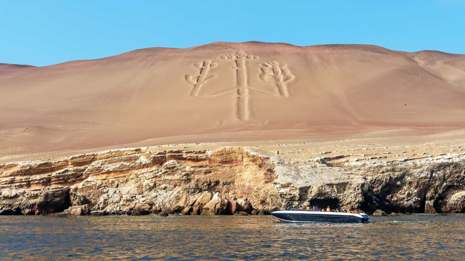 A wide shot of the Paracas Candelabra geoglyph in Peru, a well-known historical landmark etched into a huge sandhill and seen from a boat on the water.