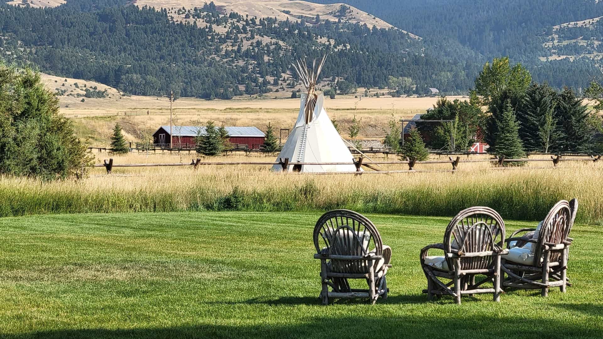 A tepee stands in a golden field in Paradise Valley, Montana, with rustic chairs in the foreground and forested hills behind.