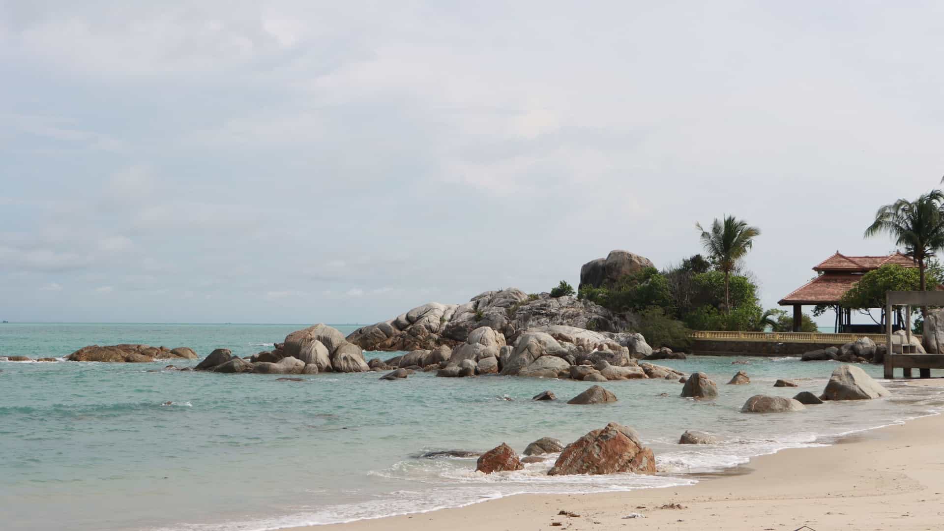 A wide shot of Parai Beach, Indonesia, with a small wooden structure with a red roof on a pier, a sandy beach, and large granite boulders in the water.