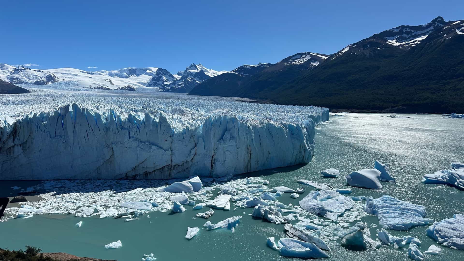 A massive, bright blue and white glacier face meets an icy, turquoise lake, with snow-capped mountains in the background.