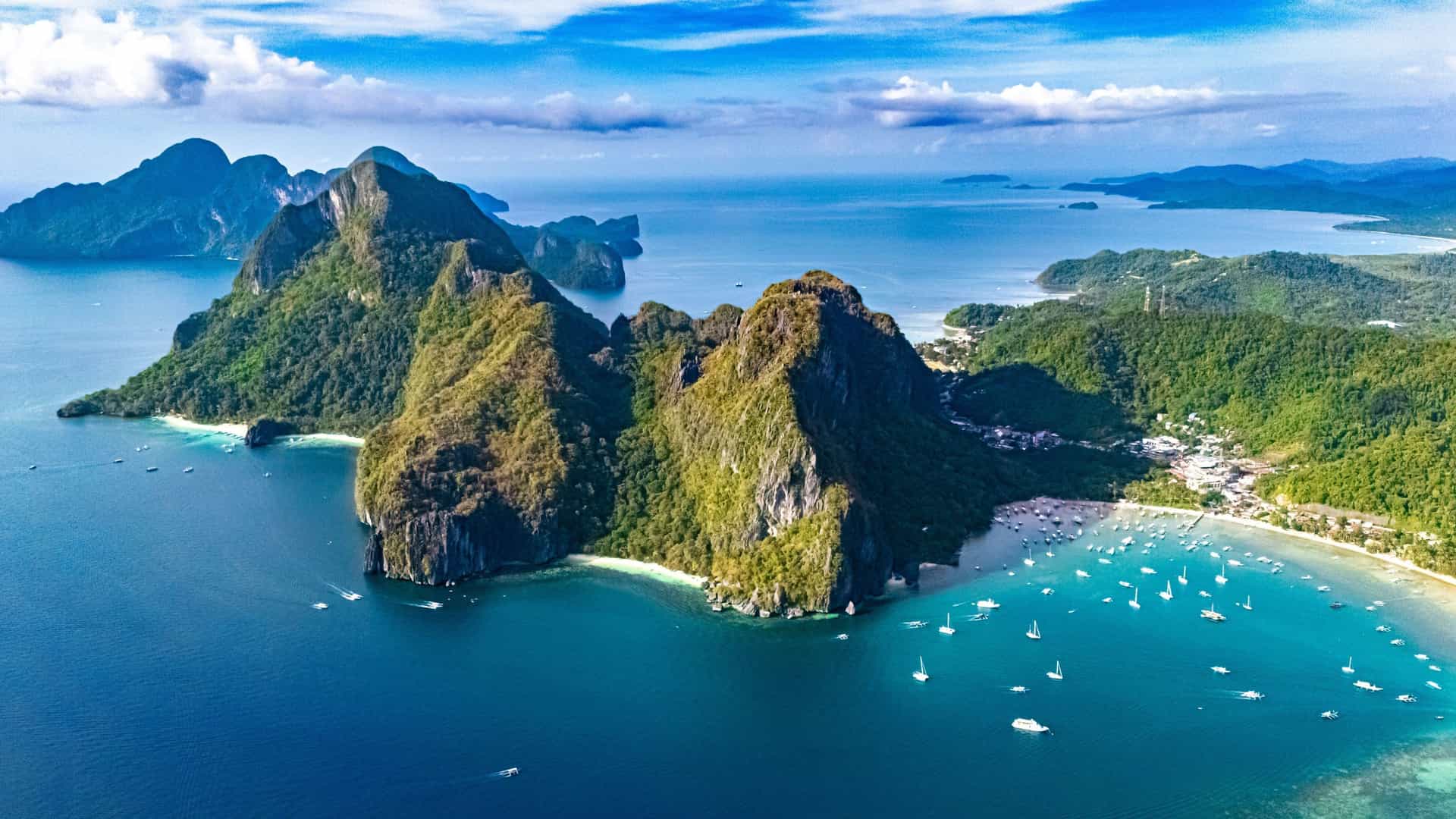 An aerial panoramic view of El Nido, Palawan, in the Philippine Sea, with large, lush mountains surrounded by a clear turquoise bay full of boats.