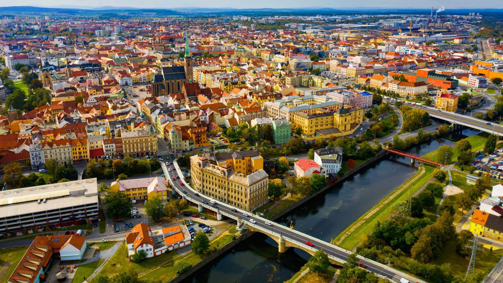 An aerial panoramic shot of the beautiful city of Plzeň, Czech Republic, with colorful buildings, a river, and bridges under a blue sky.