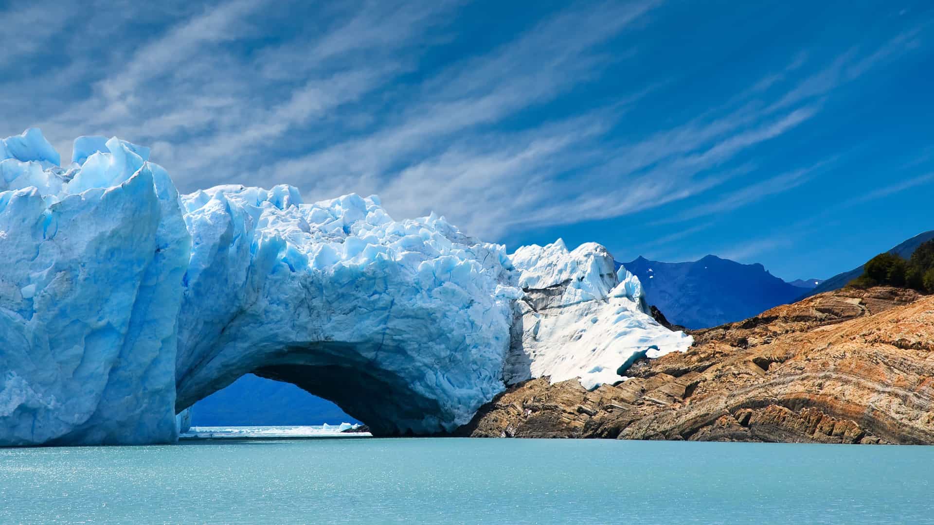 A breathtaking view of the massive Pio XI Glacier in Chile, showing its bright blue ice arching over the water, with a rocky shoreline and mountains in the distance under a blue sky.