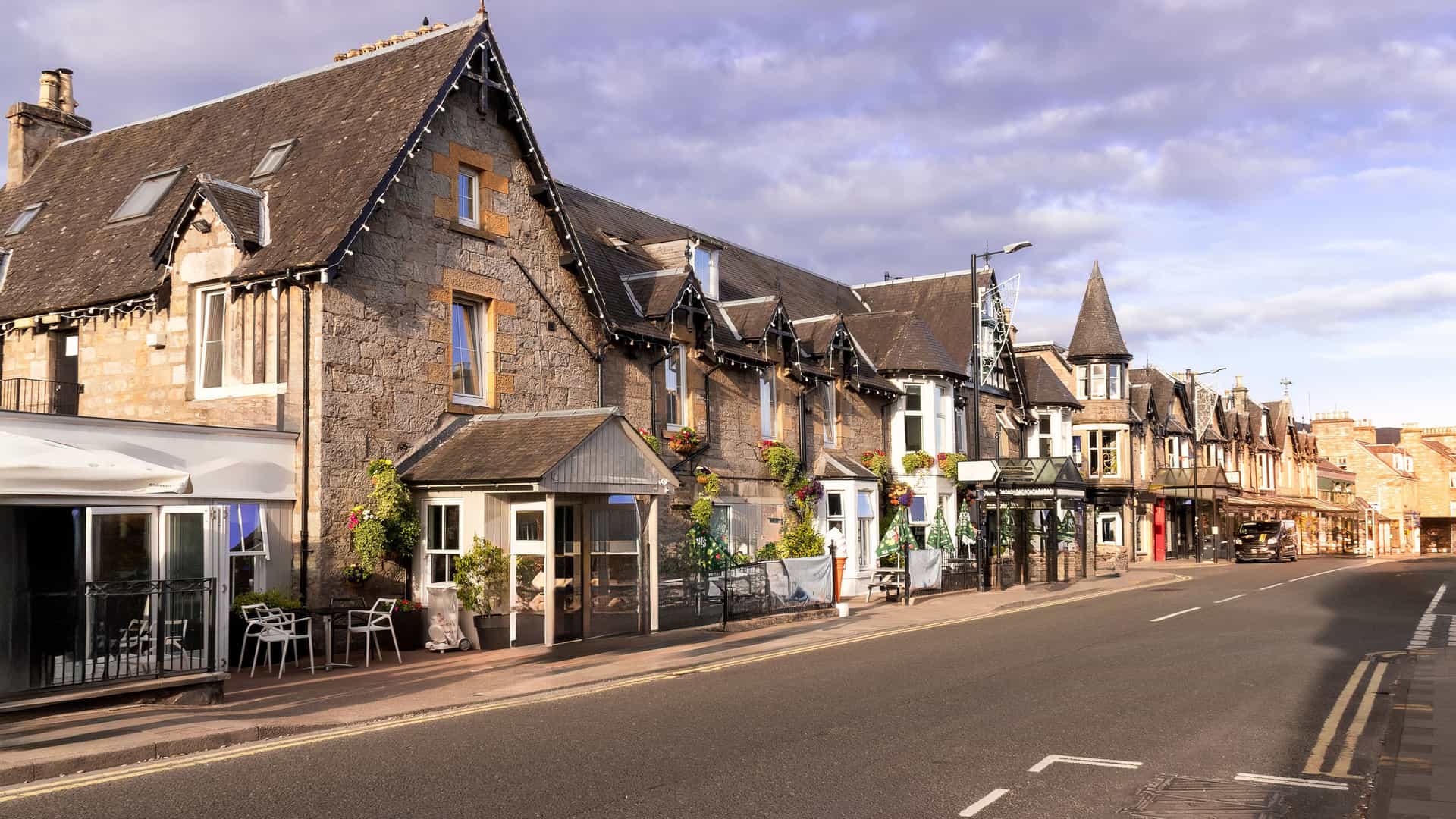 A horizontal view of a street in Pitlochry, Scotland, lined with charming, traditional stone buildings. A paved road with double yellow lines runs along the row of hotels and shops, leading into the distance under a partly cloudy sky.