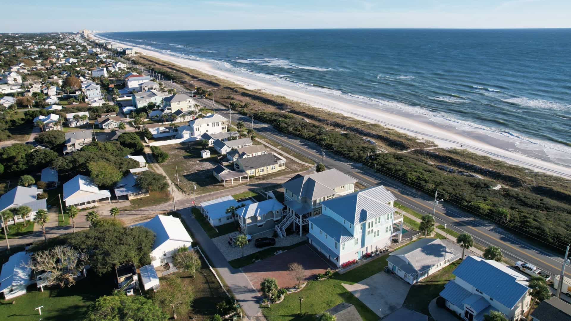 An aerial view of a beachfront town shows colorful houses next to a white-sand beach and the blue ocean.