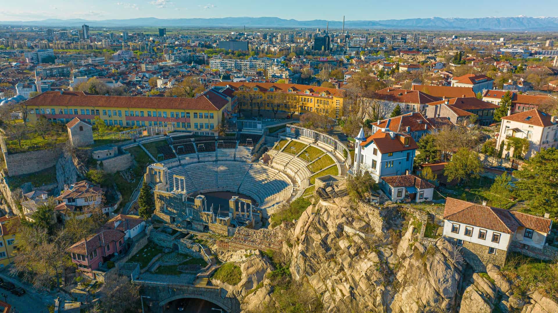 An aerial view of the ancient Roman Theatre of Philippopolis in Plovdiv, Bulgaria, with stone seating and a stage in the foreground and the modern city skyline and mountains in the background.