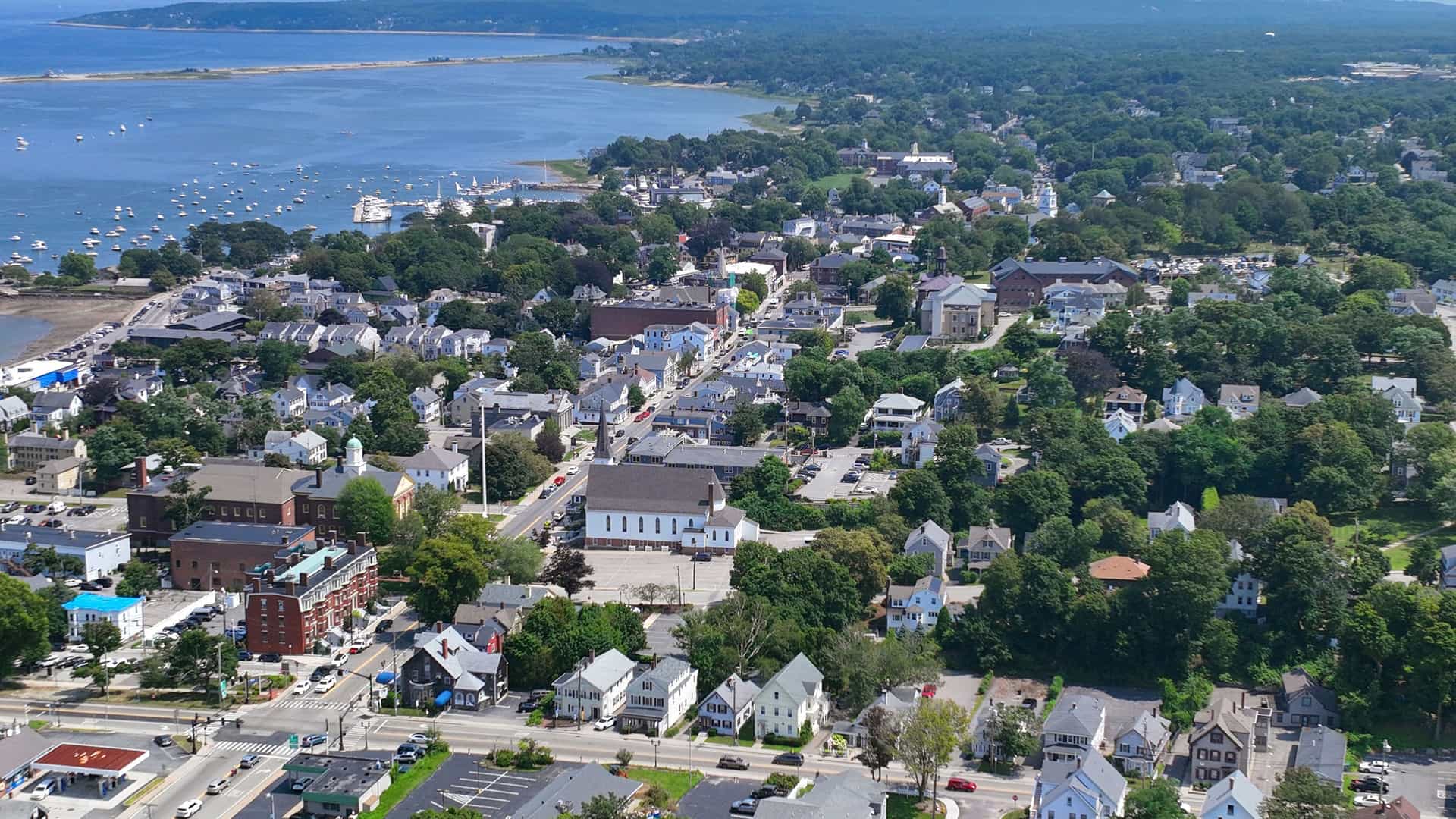 An aerial view of Plymouth, Massachusetts, showing the town's residential and commercial buildings nestled among lush green trees. A large harbor filled with many small boats is visible on the left, with the main body of water and a distant shoreline stretching out under a bright, sunny sky.