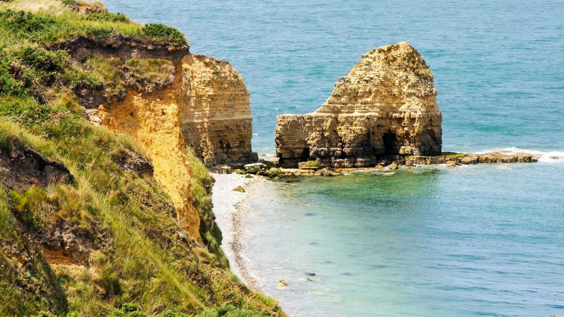 The historic Pointe du Hoc coastline shows sheer, grassy, tan cliffs meeting a small shingle beach and the clear, turquoise water.