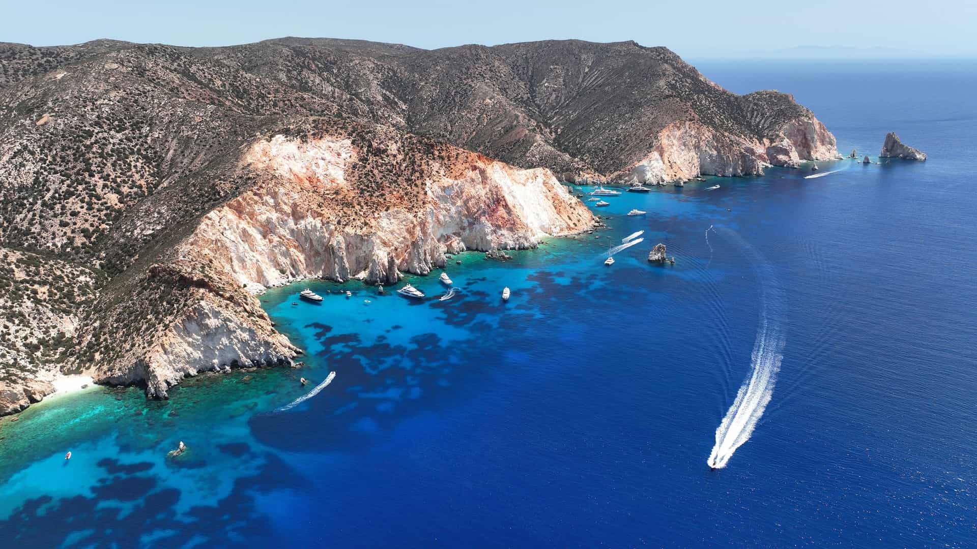 An aerial view of the rugged coastline of Poliegos, Greece, with steep, rocky cliffs meeting the clear turquoise and deep blue waters of the Aegean Sea. Several small boats are anchored in coves, and a speedboat is visible in the foreground, leaving a white wake behind it.