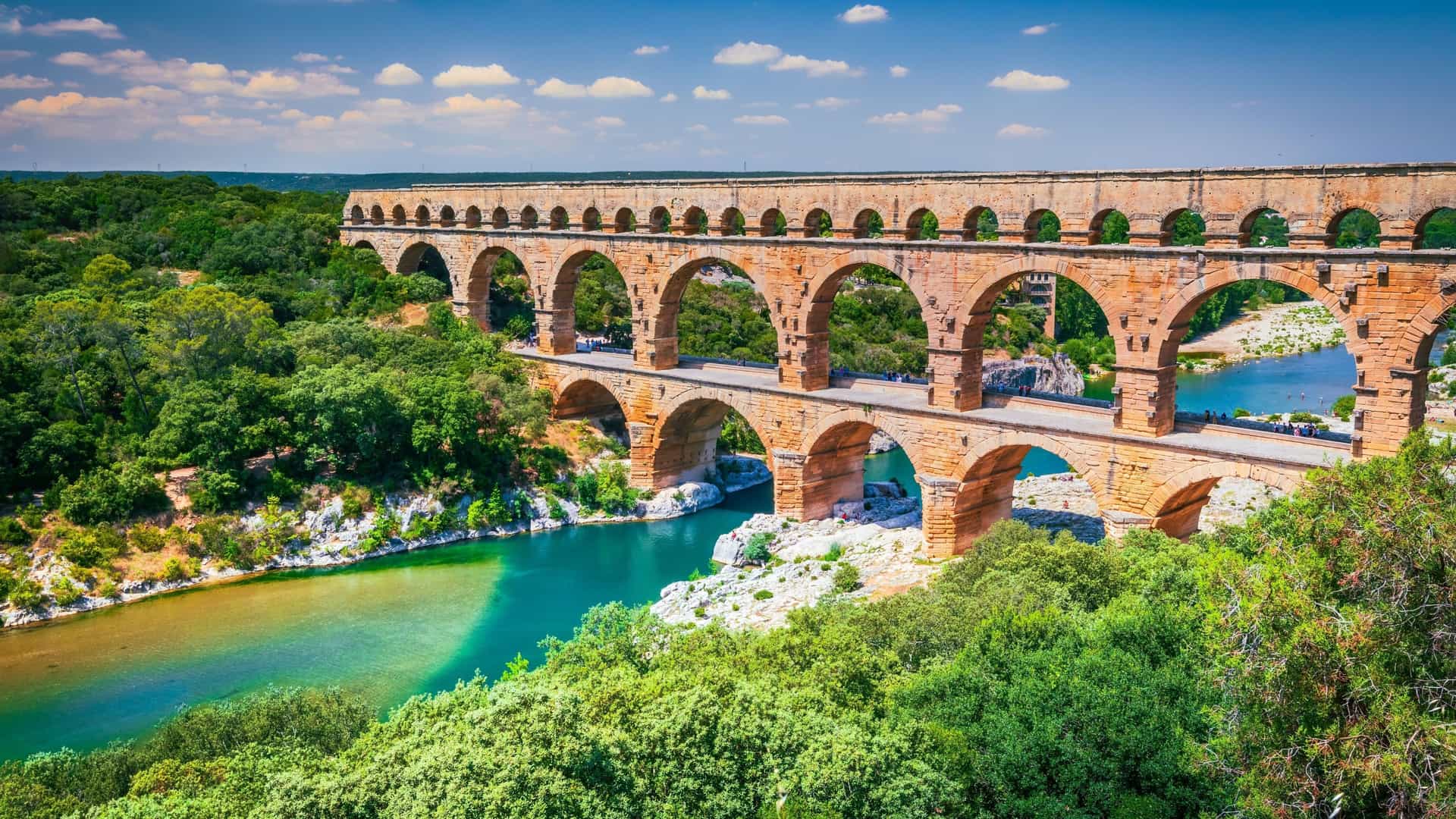 A panoramic view of the ancient Roman aqueduct, Pont du Gard, in France, showing its three tiers of arches spanning the beautiful, teal-colored Gardon River and surrounded by green trees.