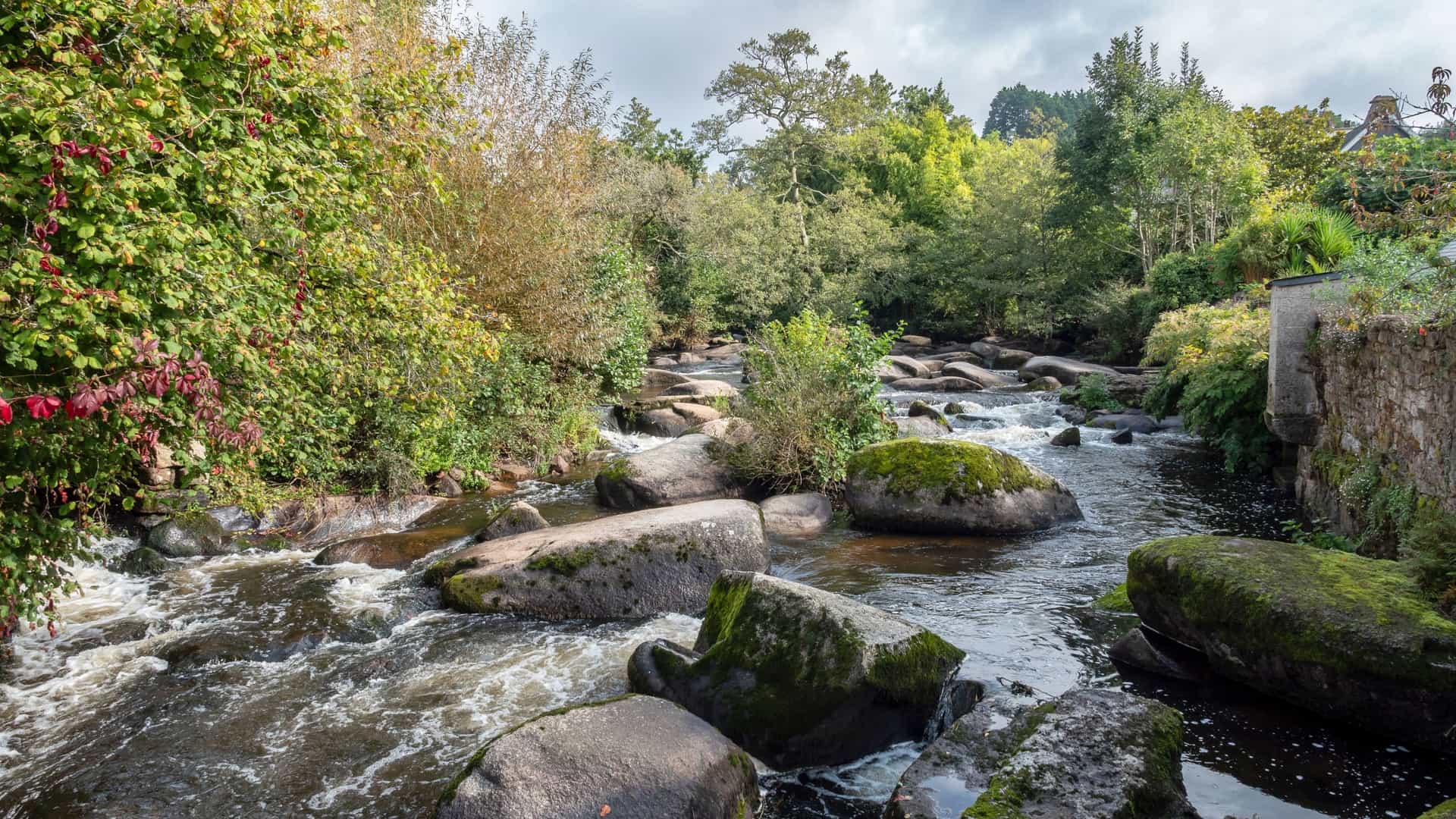 A picturesque view of the Aven River flowing through Pont-Aven, France, with large moss-covered boulders in the shallow, rushing water and trees on the banks.