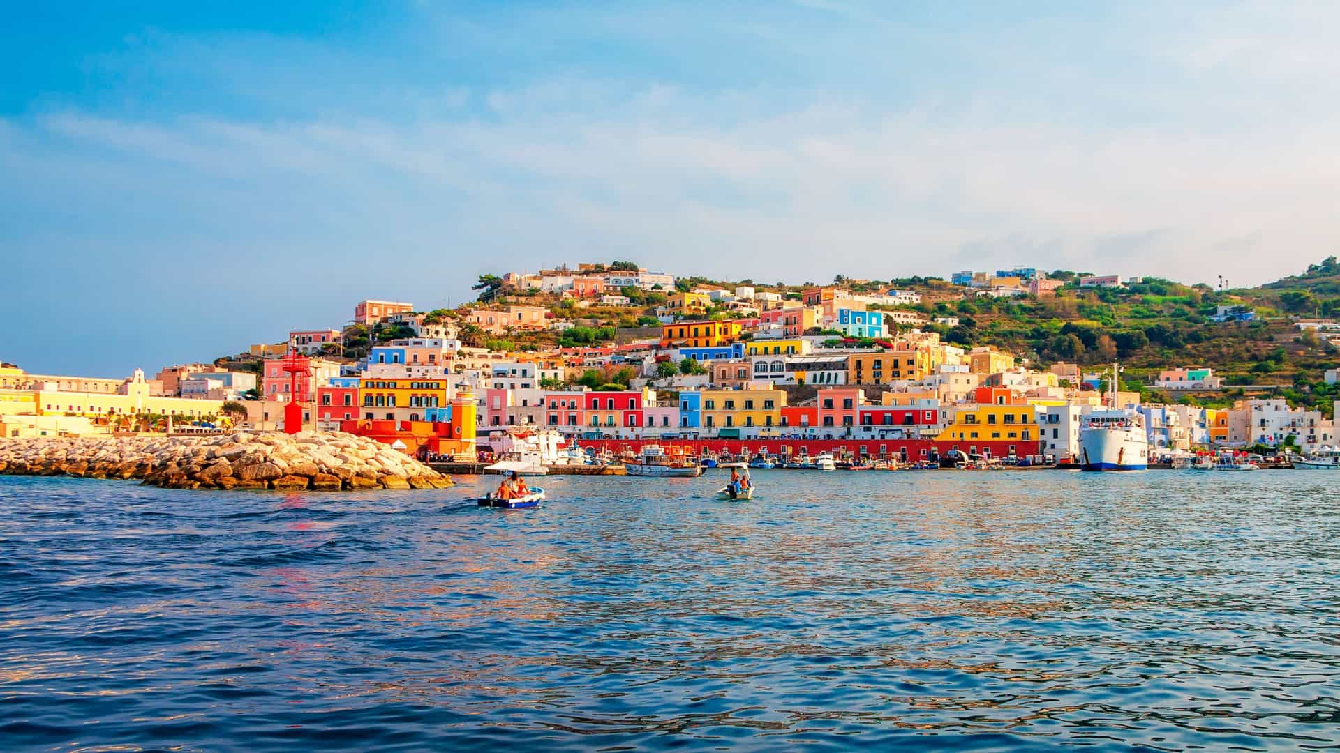 A colorful view of the port of Ponza Island in Italy, with brightly painted houses stacked on the hillside overlooking the blue Tyrrhenian Sea and boats docked along the waterfront.
