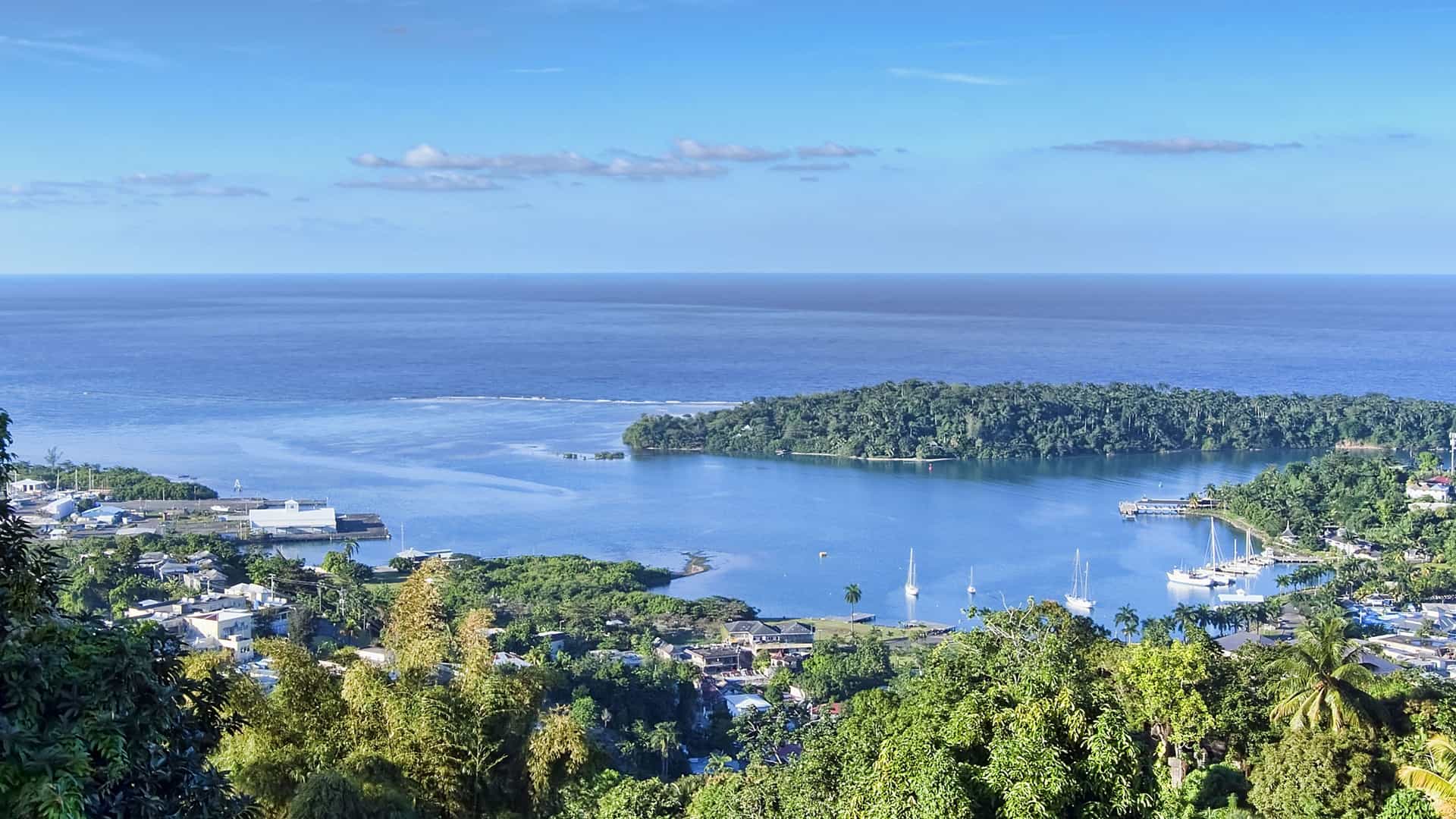 A high-angle panoramic view of the Port Antonio harbor in Jamaica, with the beautiful blue Caribbean Sea and a lush, tree-covered peninsula in the foreground and boats anchored in the bay.