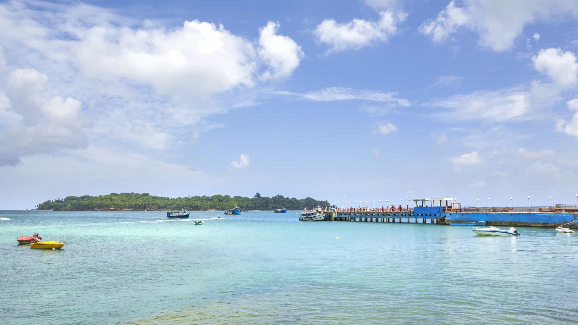 A serene view of the turquoise waters of the Andaman Sea, with a long blue pier stretching out from Port Blair, and small boats scattered on the water, with Ross Island in the distance.