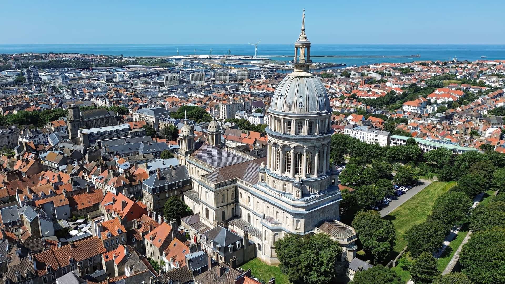 An aerial view of the historic city of Boulogne-sur-Mer, France, showcasing the impressive Basilique Notre-Dame de Boulogne and the surrounding terracotta rooftops, with the port of Calais and the English Channel in the distance.