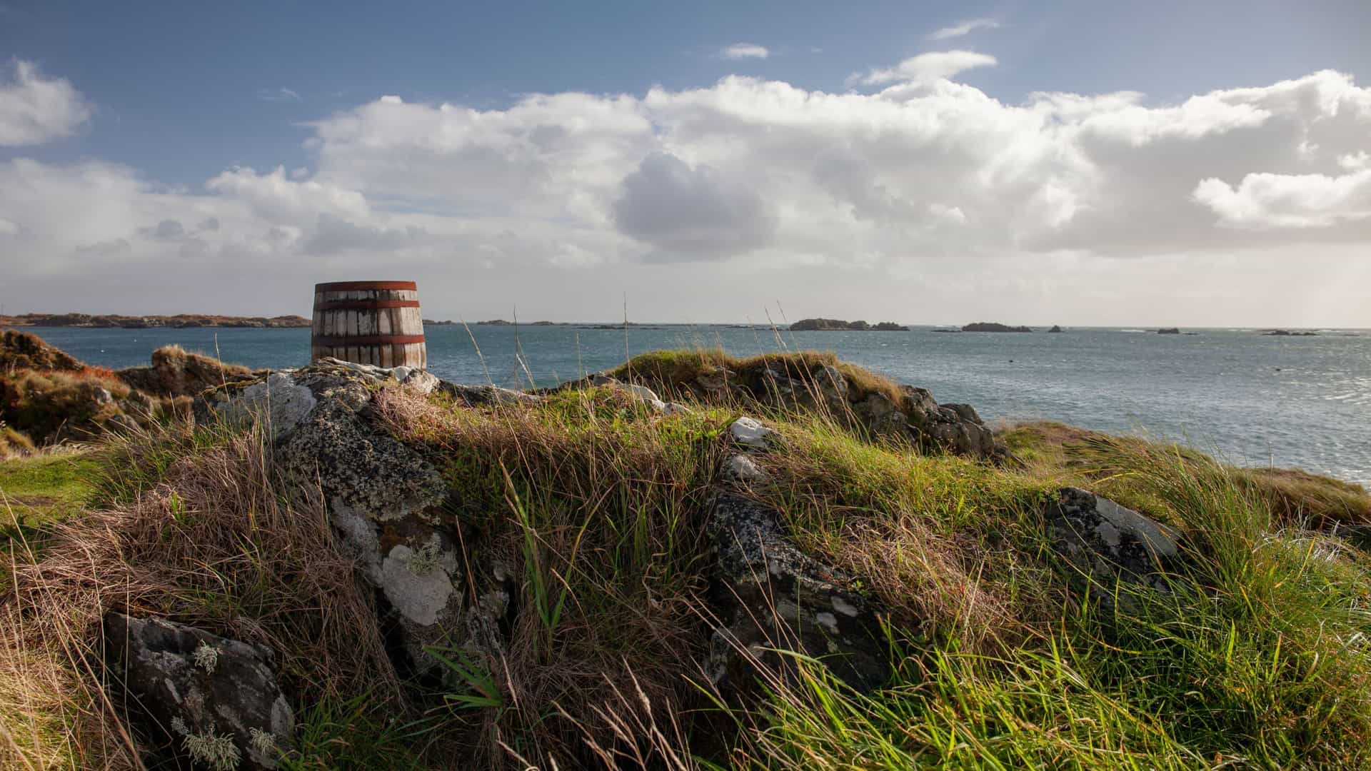A grassy and rocky hillside overlooking the sea on the Isle of Islay, Scotland, with a large, rusted metal barrel monument in the background.