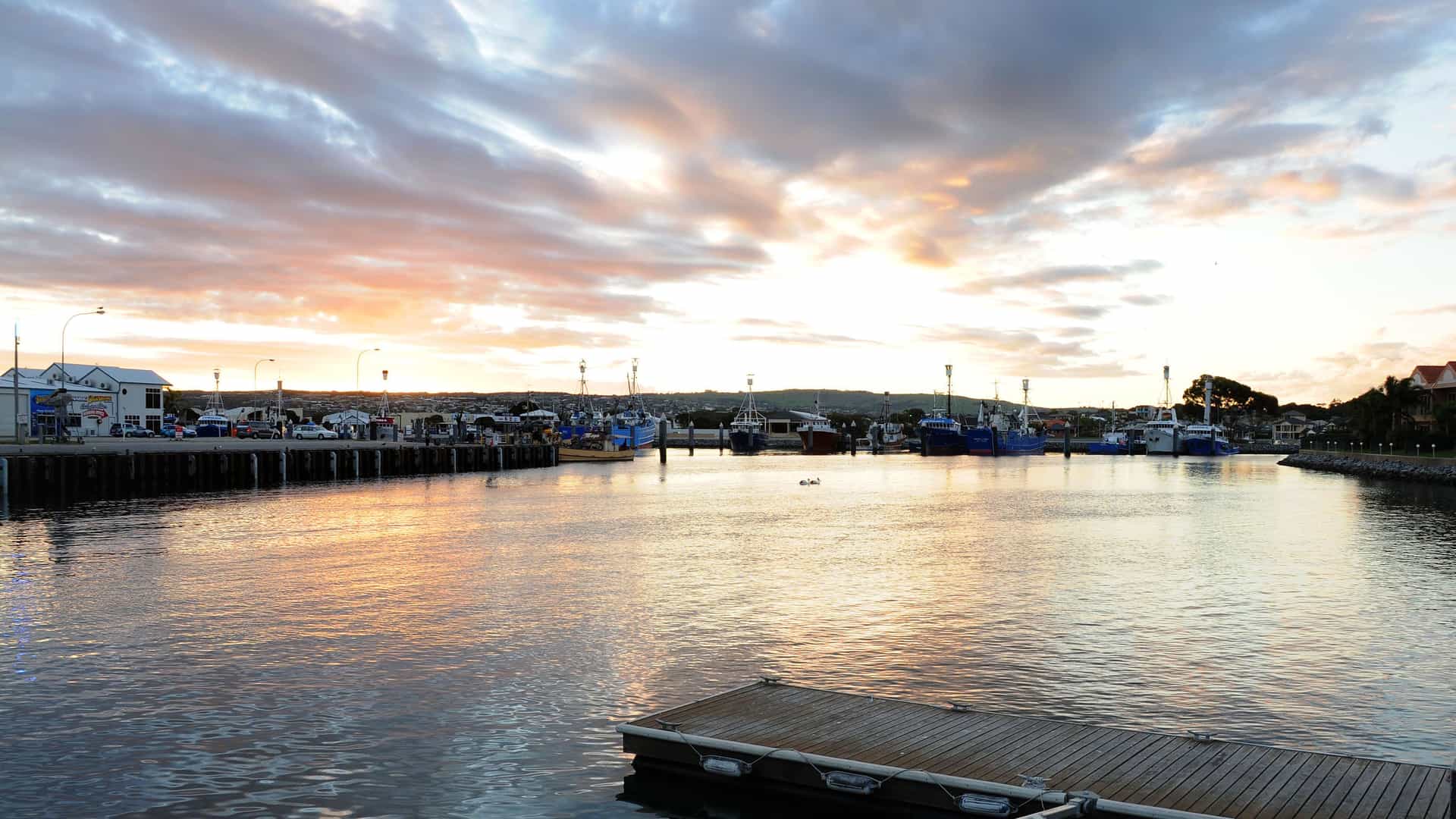 A calm harbor in Port Lincoln, South Australia, at sunrise, with a variety of fishing boats and yachts docked at the marina and the town in the distance under a dramatic, cloudy sky.
