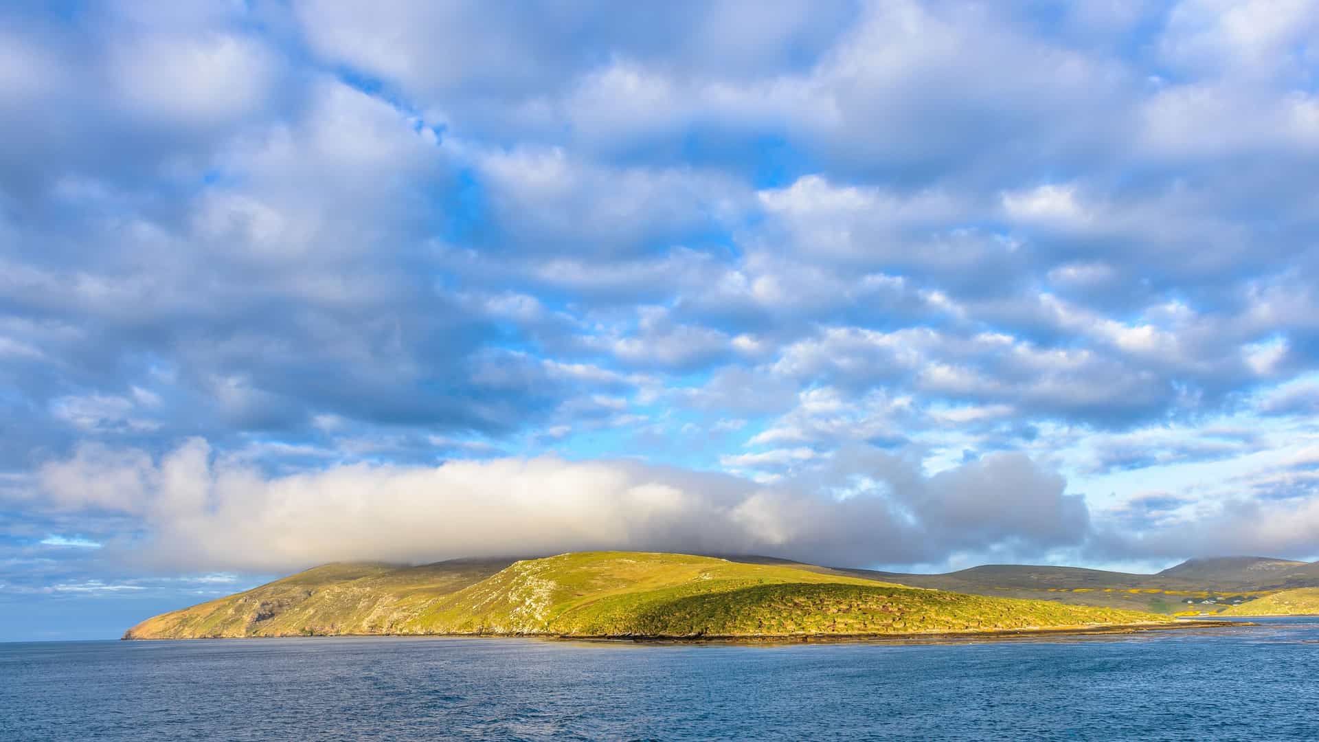 A panoramic view of the rugged, green landscape of the Falkland Islands, with a low cloud covering the top of a hill and the blue sea in the foreground under a dramatic sky.