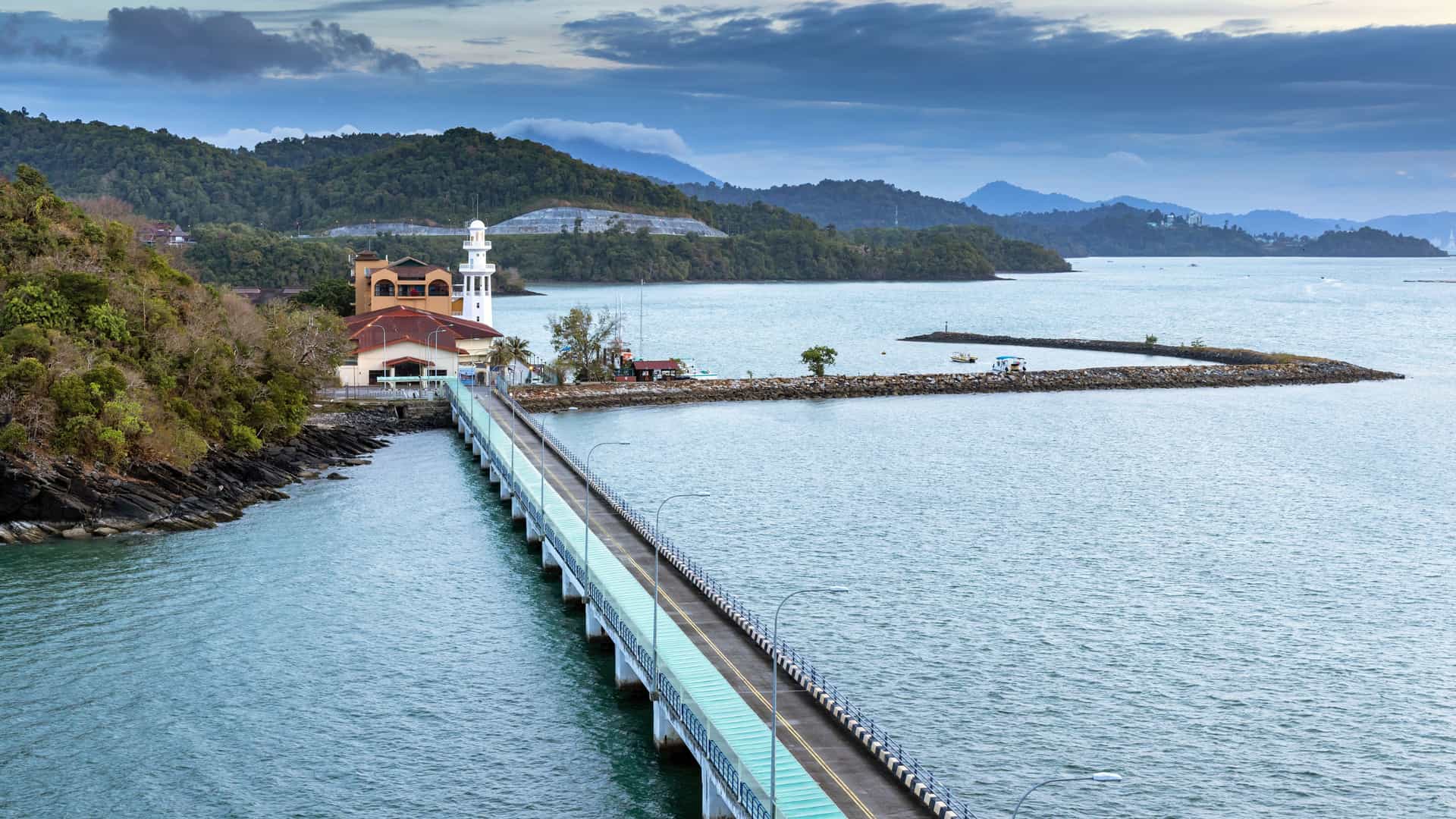 A peaceful coastal view in Porto Malai with a long pier stretching over calm water toward a cluster of buildings topped with red and beige roofs, including a white lighthouse, all set against forested hills and distant mountains beneath a cloudy sky.