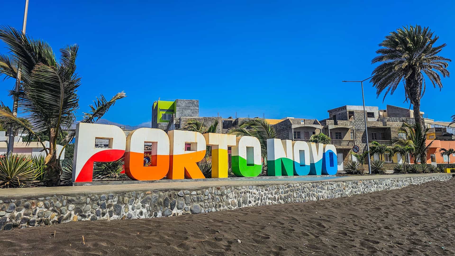 A large, colorful "PORTO NOVO" sign stands prominently on the beachfront, framed by palm trees and buildings, welcoming visitors to the city in Cape Verde.