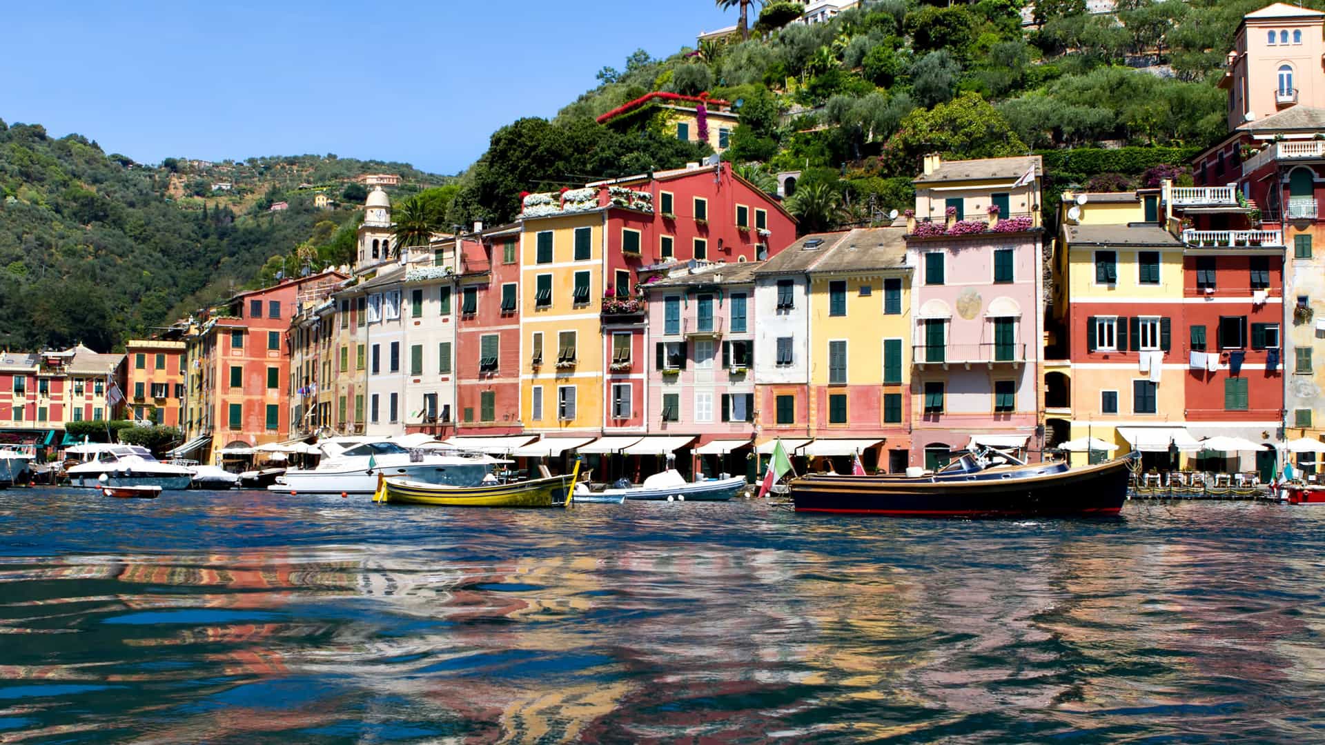 A classic view of the picturesque harbor of Portofino, Italy, with its iconic, brightly painted buildings reflected on the shimmering blue water and lush green hills in the background.