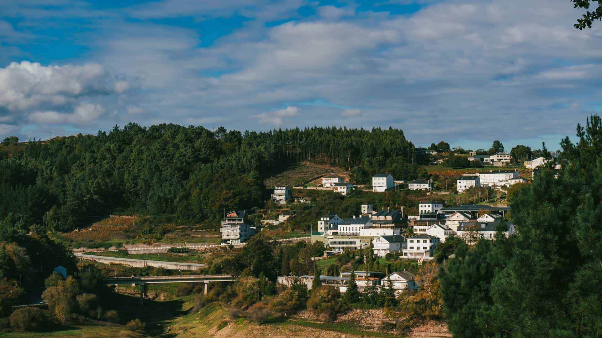 A scenic view of the town of Portomarín, Spain, with its white buildings and houses built into a lush green, forested hillside. In the foreground, a bridge crosses a dry, wide riverbed under a sky with scattered clouds.