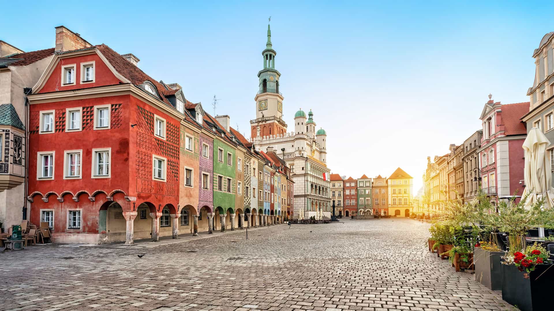 The historic Old Market Square in Poznań, Poland, with its cobblestone pavement. The left side of the square is lined with a row of colorful, narrow merchant houses, while the ornate Poznań Town Hall, with its tall clock tower, dominates the center. The warm light of the rising or setting sun casts a golden glow on the buildings.