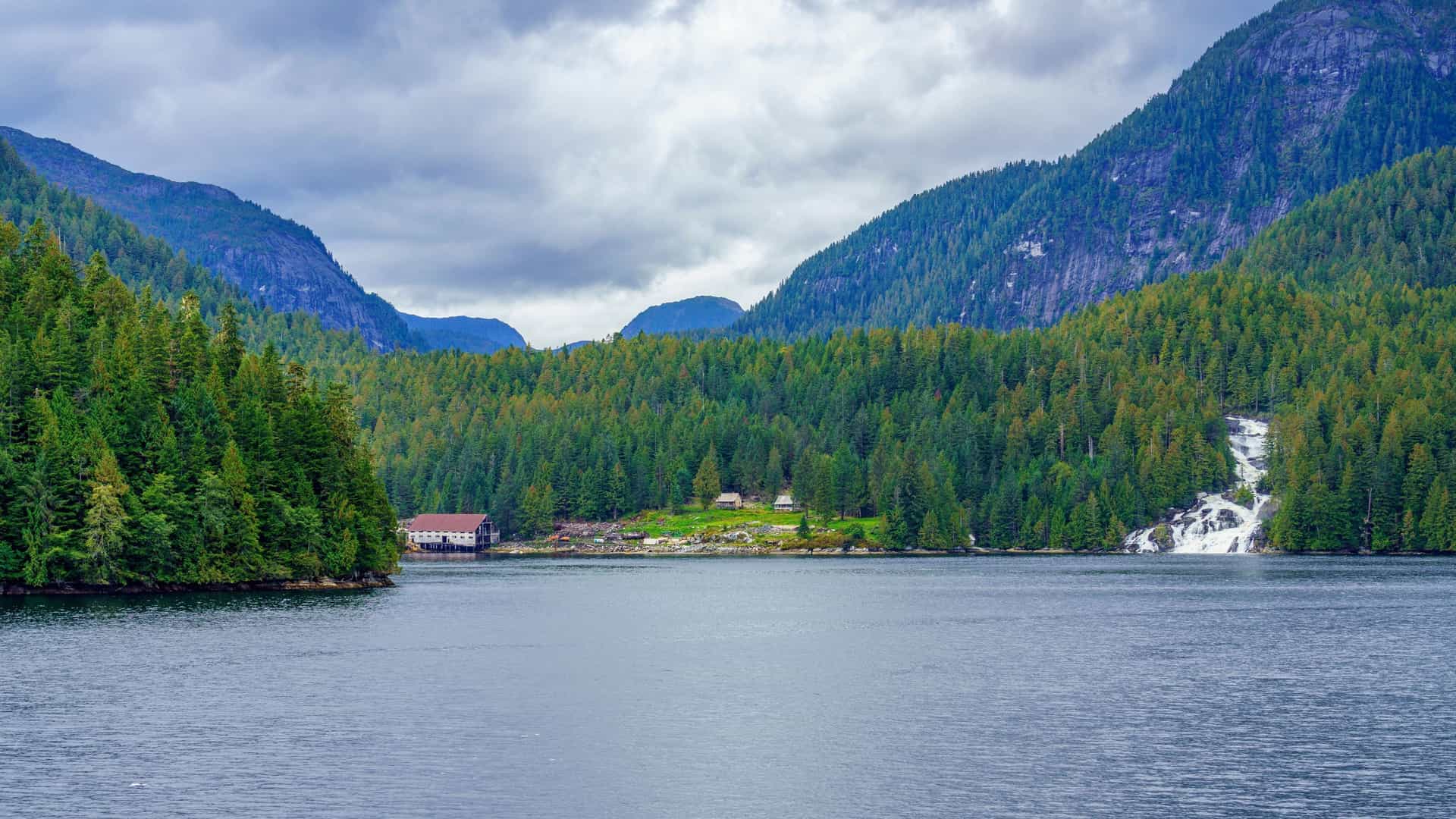 A stunning landscape shot of Princess Royal Channel, Canada, with a small house nestled among dense green forests and a waterfall flowing down the hillside into the tranquil water.