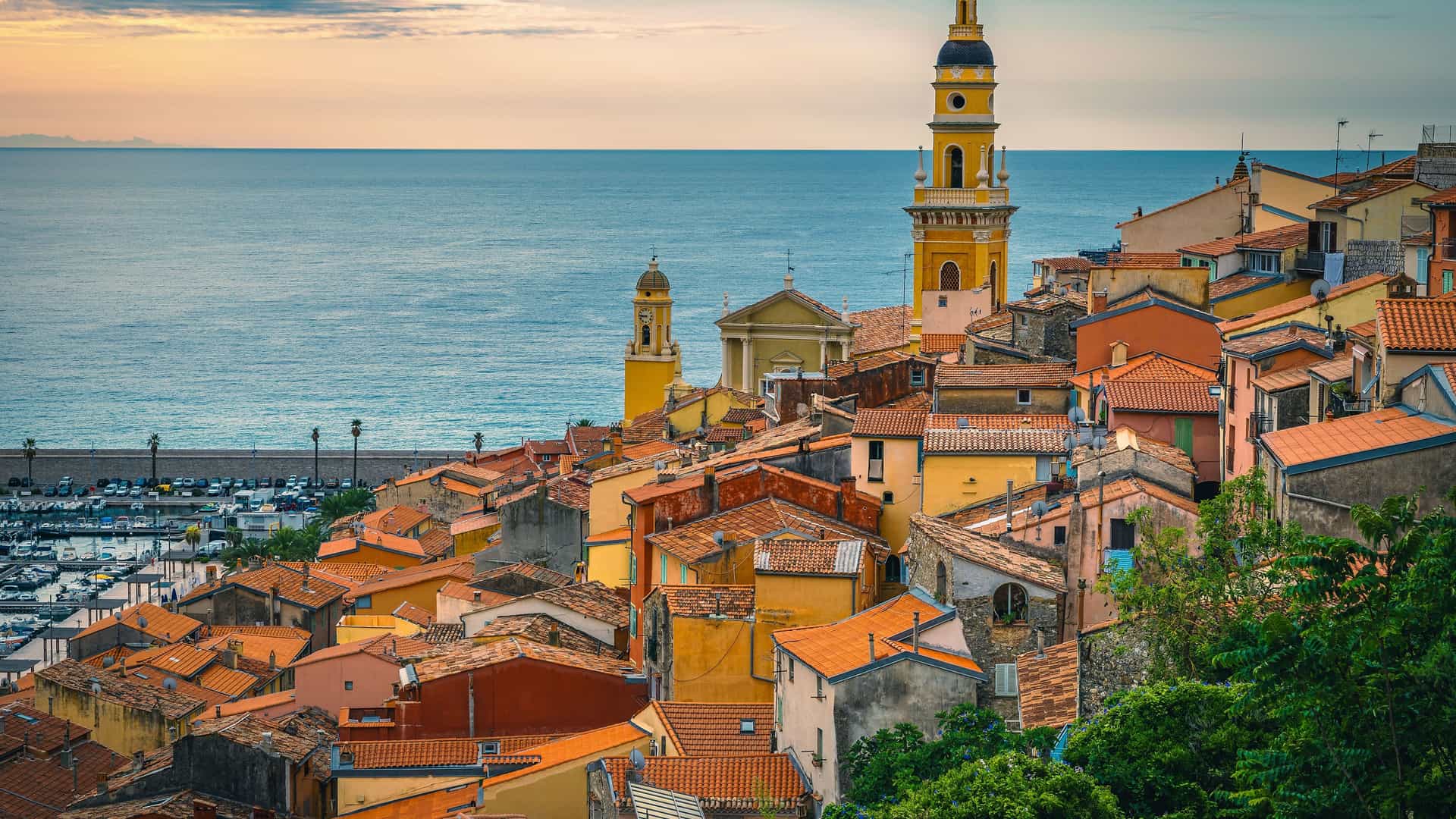 A scenic view of a coastal town in Provence, France, with a prominent yellow bell tower rising from a cluster of buildings with terracotta roofs. The town is built on a hillside that slopes down to a marina filled with boats, all set against the blue Mediterranean Sea and a hazy sky.