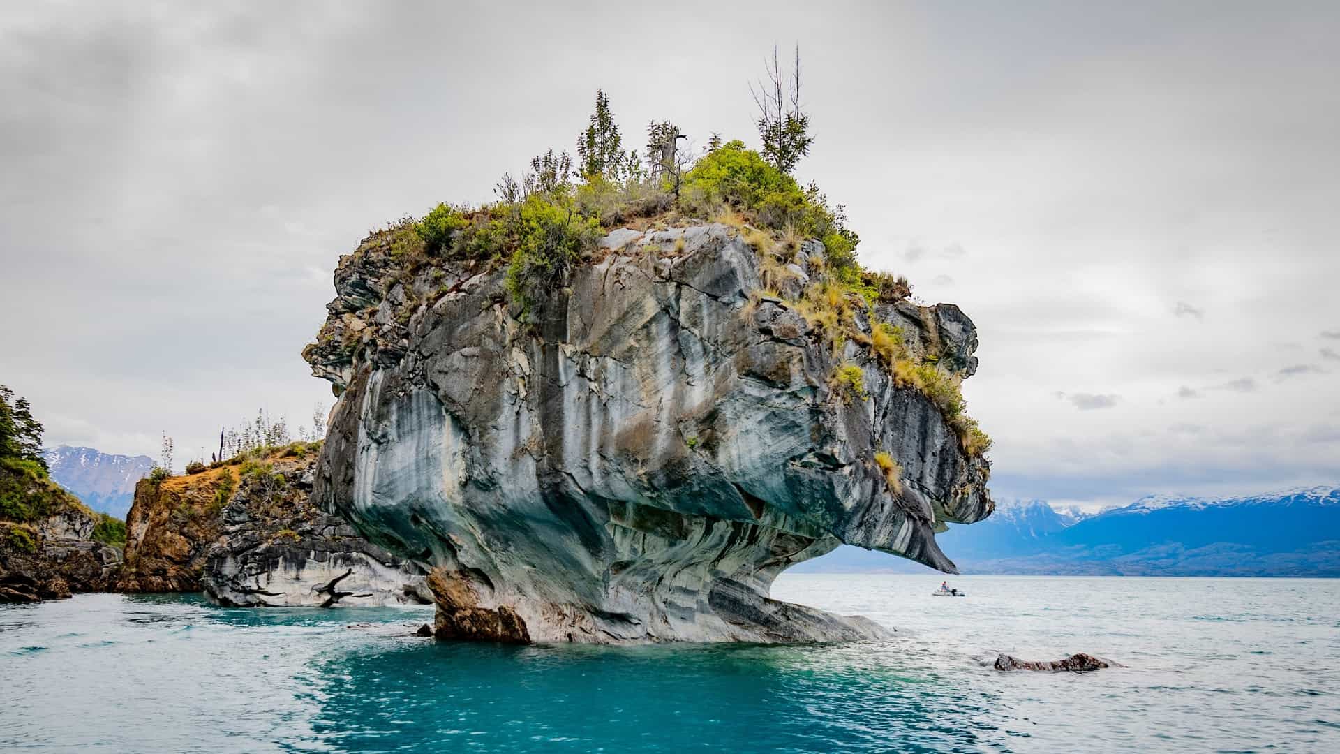 A unique, undercut marble cave formation, resembling a small island with trees, rises out of a turquoise lake under a gray sky.