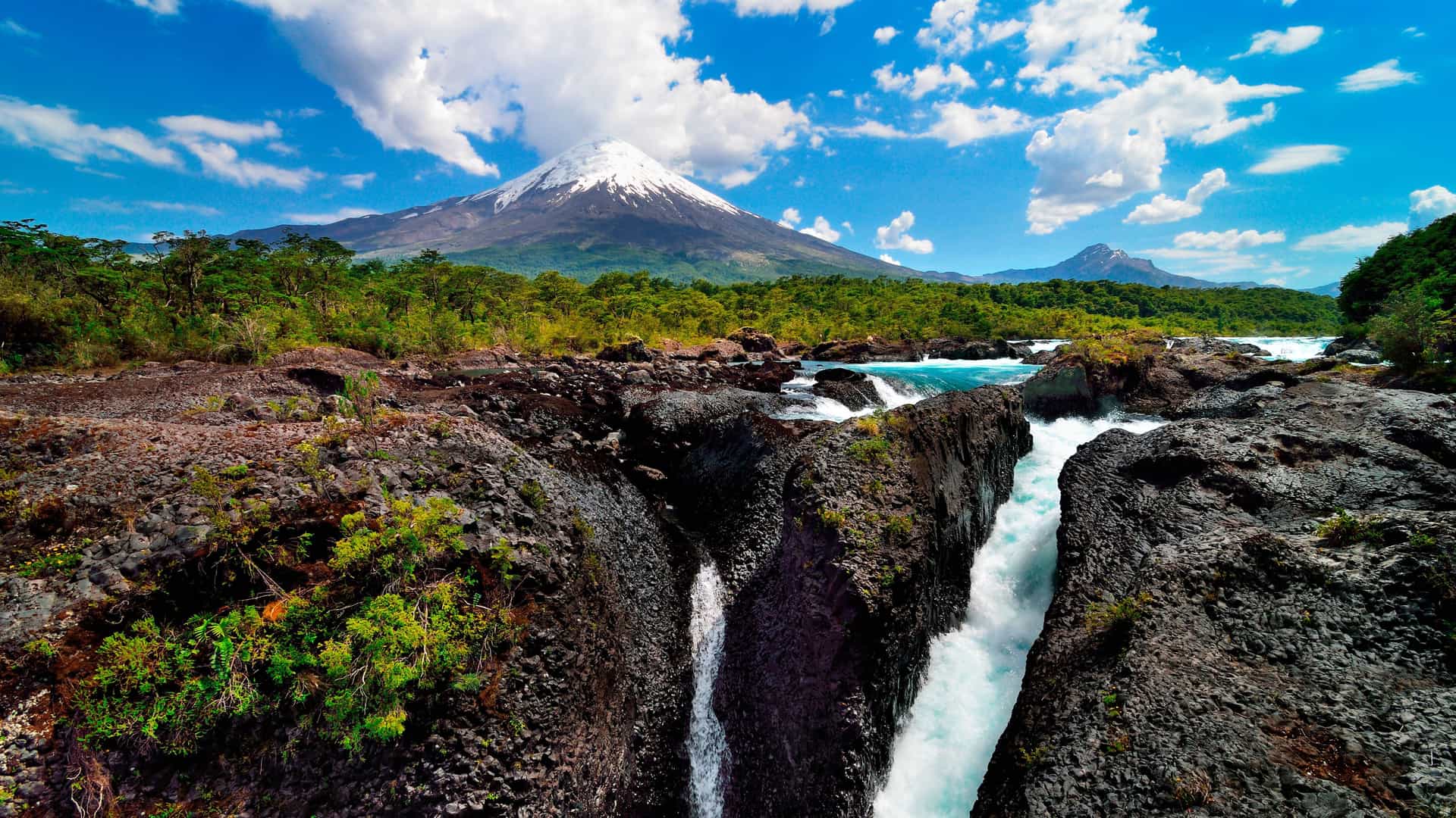 A wide shot of the spectacular Petrohué Falls near Puerto Montt, Chile, with turquoise water cascading over volcanic rock, set against the backdrop of the snow-capped Osorno Volcano.