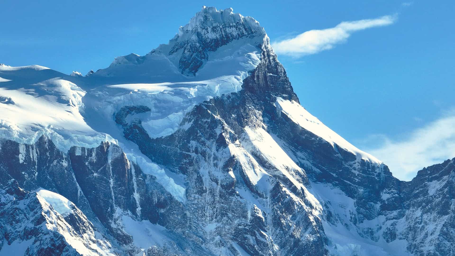 A close-up view of a massive, jagged mountain peak near Puerto Natales, Chile, covered in snow and glaciers. The steep, rocky slopes are dusted with a thick layer of white, contrasting sharply with the deep blue sky.