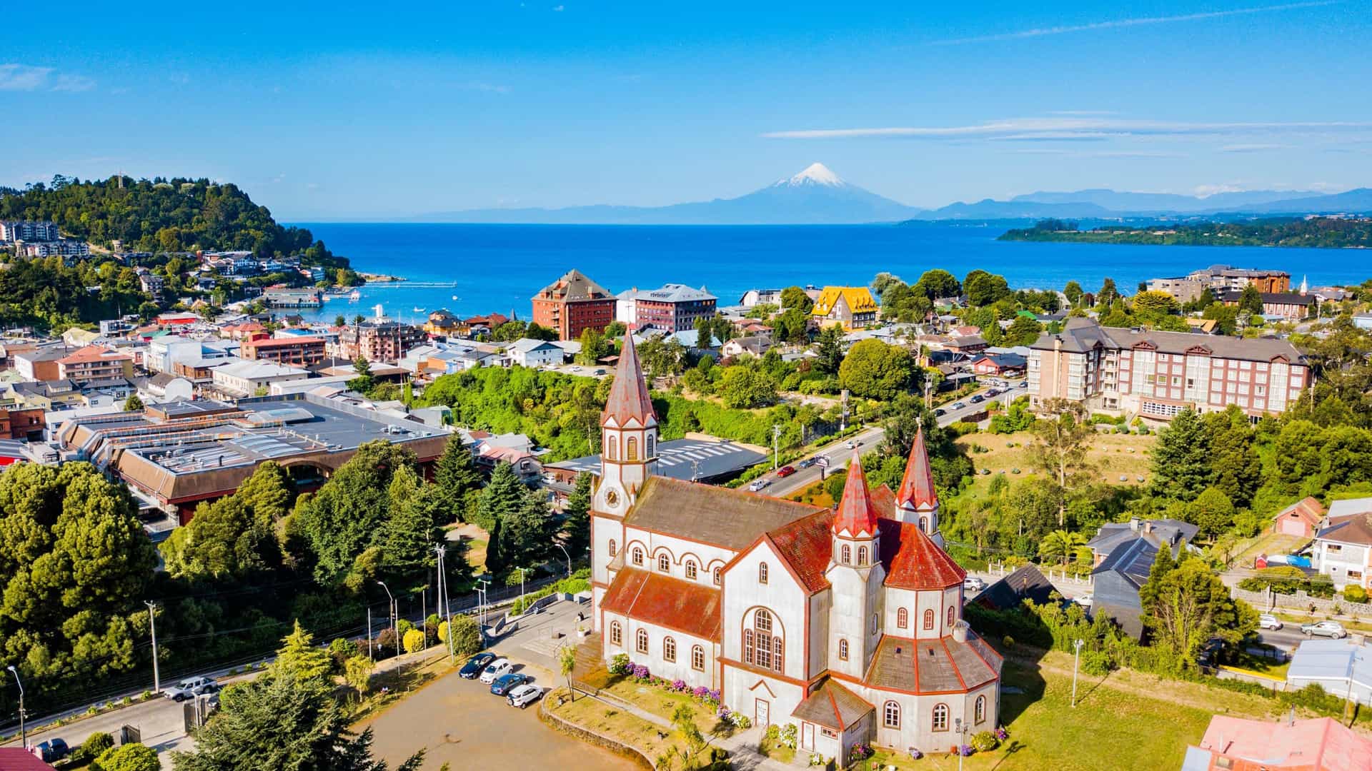 An aerial view of the town of Puerto Varas, Chile, with a prominent red-roofed church in the foreground. The town sits on the shore of a large blue lake, with the snow-capped Osorno Volcano visible in the distance across the water. The sky is bright and clear.