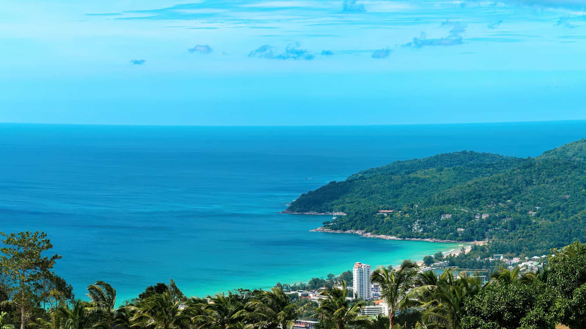 A stunning high-angle shot from a viewpoint overlooking Nai Harn Beach in Phuket, Thailand, showing a beautiful bay with turquoise water and lush green hills stretching into the deep blue ocean.