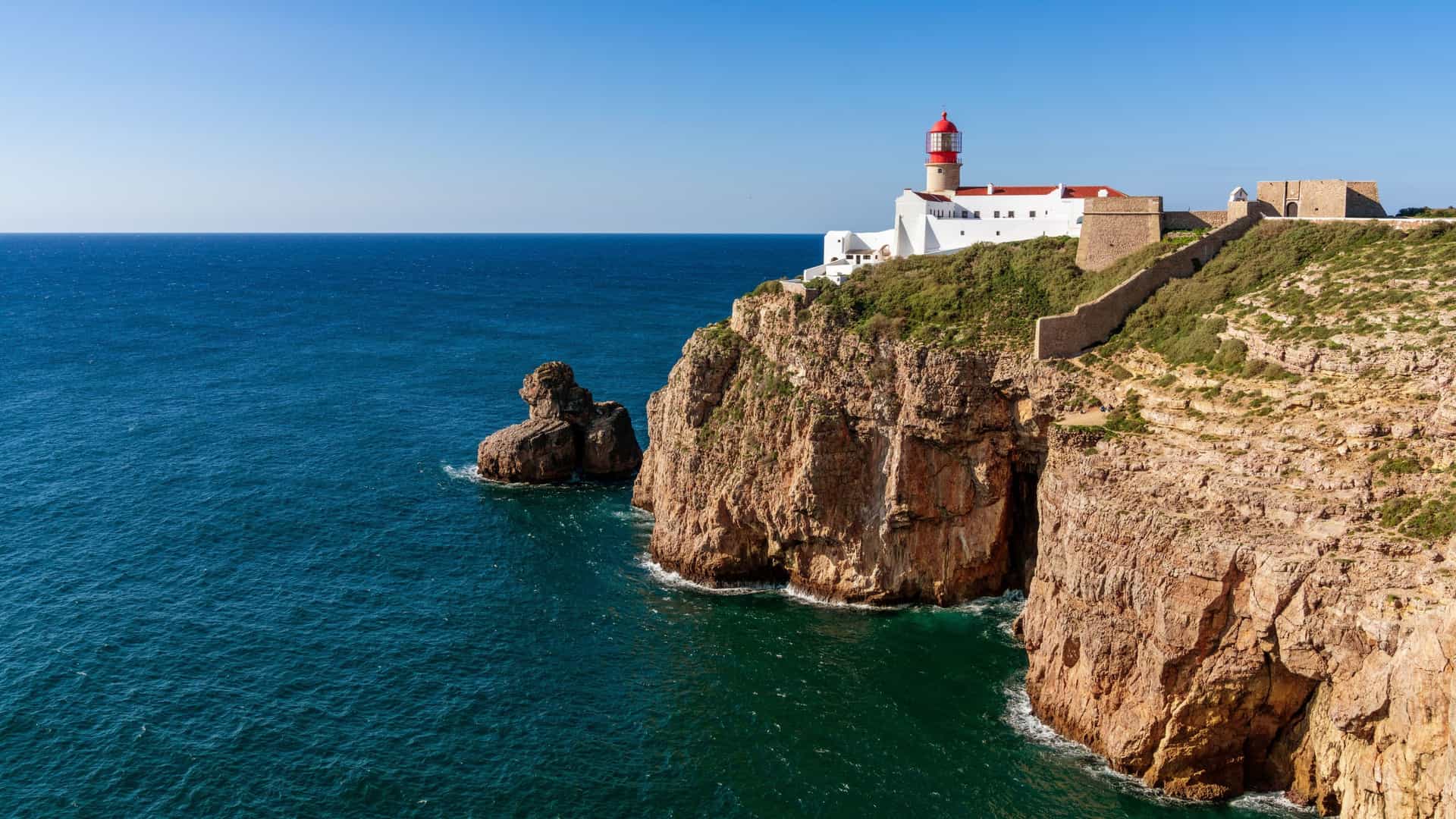 A beautiful wide shot of the Cabo de São Vicente lighthouse, a historic landmark in Portugal, perched on a dramatic cliffside overlooking the deep blue Atlantic Ocean.
