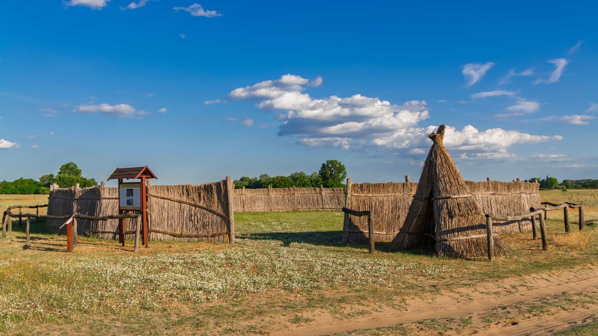 A scenic view of a traditional Puszta cultural heritage site in Hungary, featuring a rustic thatched fence and a small, pointed hut in an open field under a blue sky with scattered clouds.