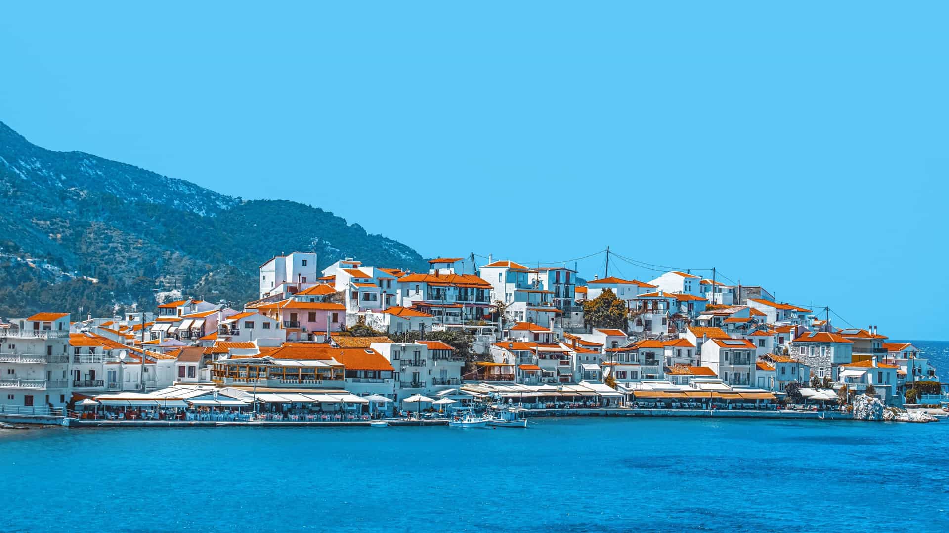 A scenic, wide-angle view of the harbor and town of Pythagoreio, Samos Island, Greece, featuring traditional white and colorful houses with red-tiled roofs built along a hillside. The foreground shows the port filled with small boats, and the clear, blue water of the Aegean Sea stretches to the horizon.