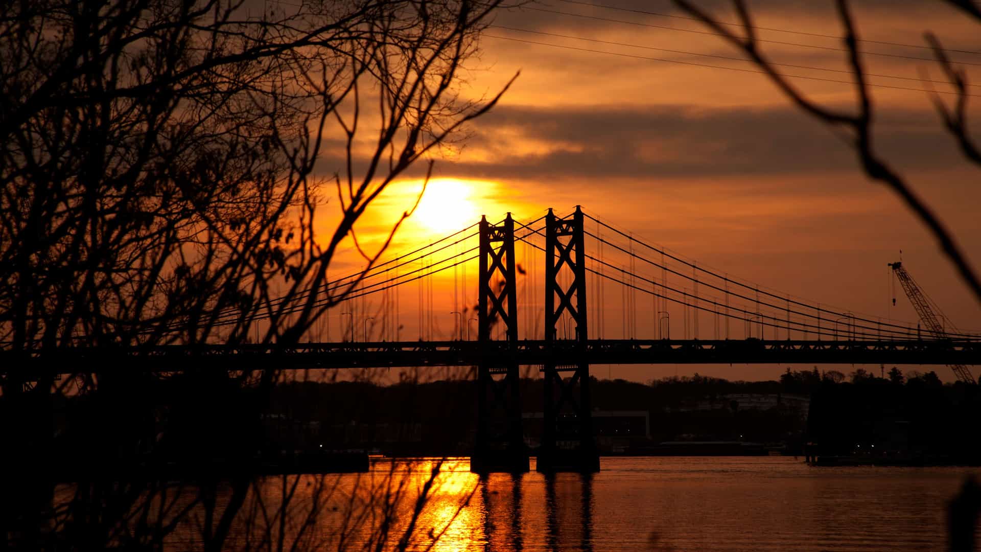A breathtaking sunset view over the Mississippi River in the Quad Cities, Iowa, with a large, silhouetted bridge framed by bare tree branches and the sun's reflection on the water.