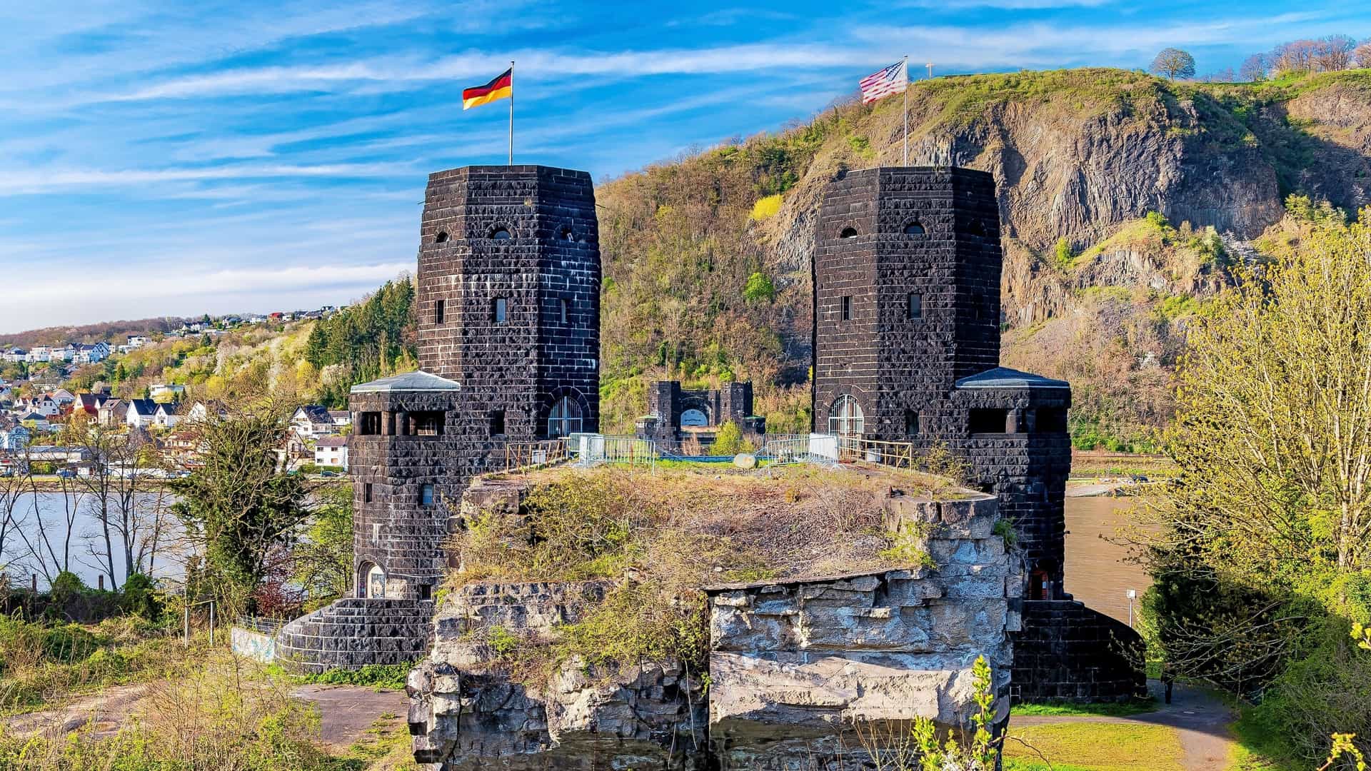 The imposing, ruined black stone towers of the Bridge at Remagen stand on a green hill, flying German and US flags.