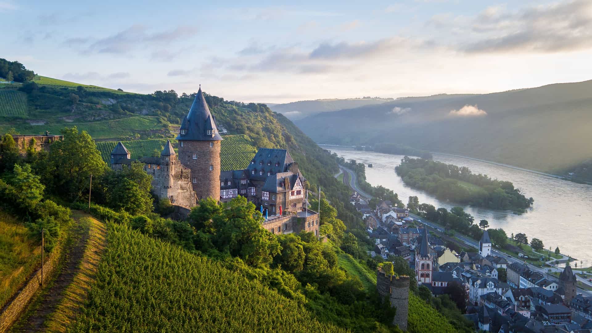 A scenic view of Stahleck Castle overlooking the town of Bacharach and the Rhine River in Germany, surrounded by vineyards and rolling green hills at sunrise.