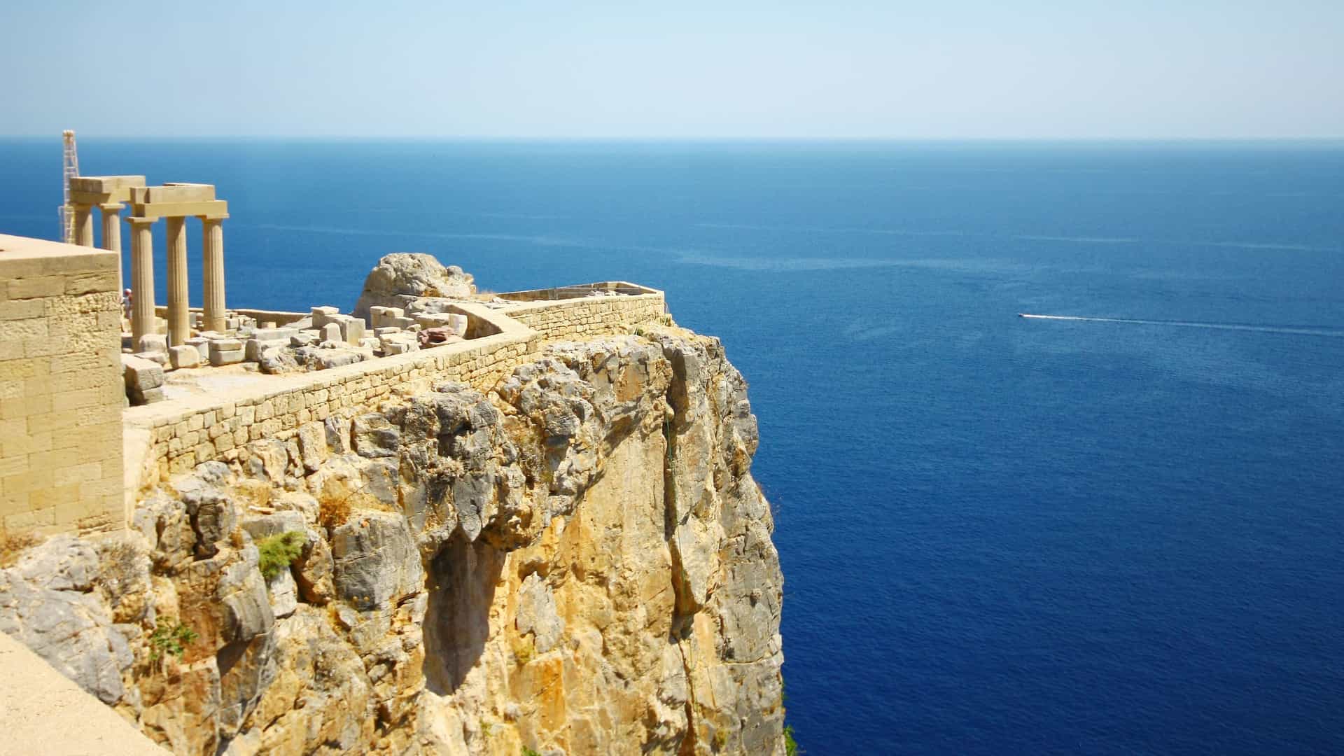 A scenic view of the Acropolis of Lindos ruins perched on a cliffside overlooking the deep blue Mediterranean Sea and a clear sky on the island of Rhodes, Greece.