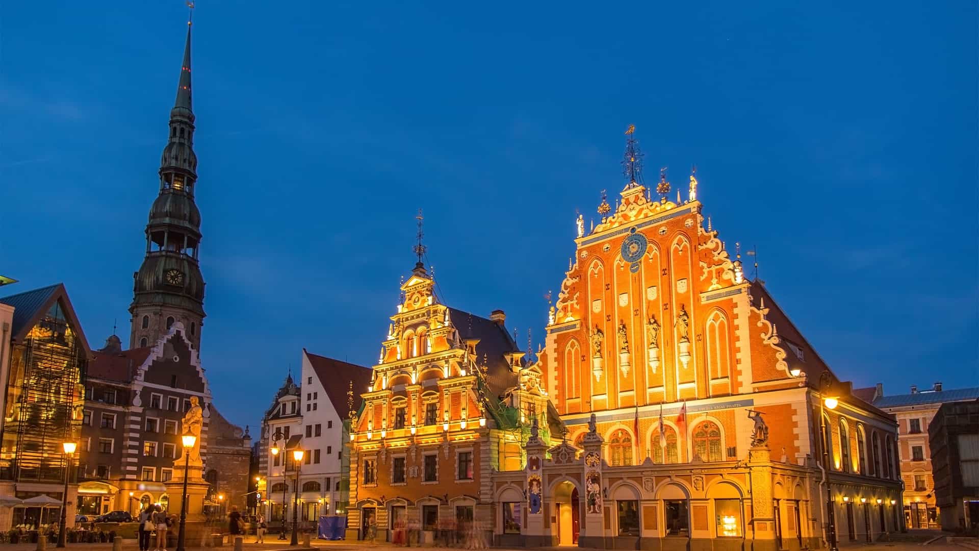 A nighttime shot of Riga's historic Old Town, featuring the beautifully illuminated House of the Blackheads and St. Peter's Church steeple against a deep blue sky.