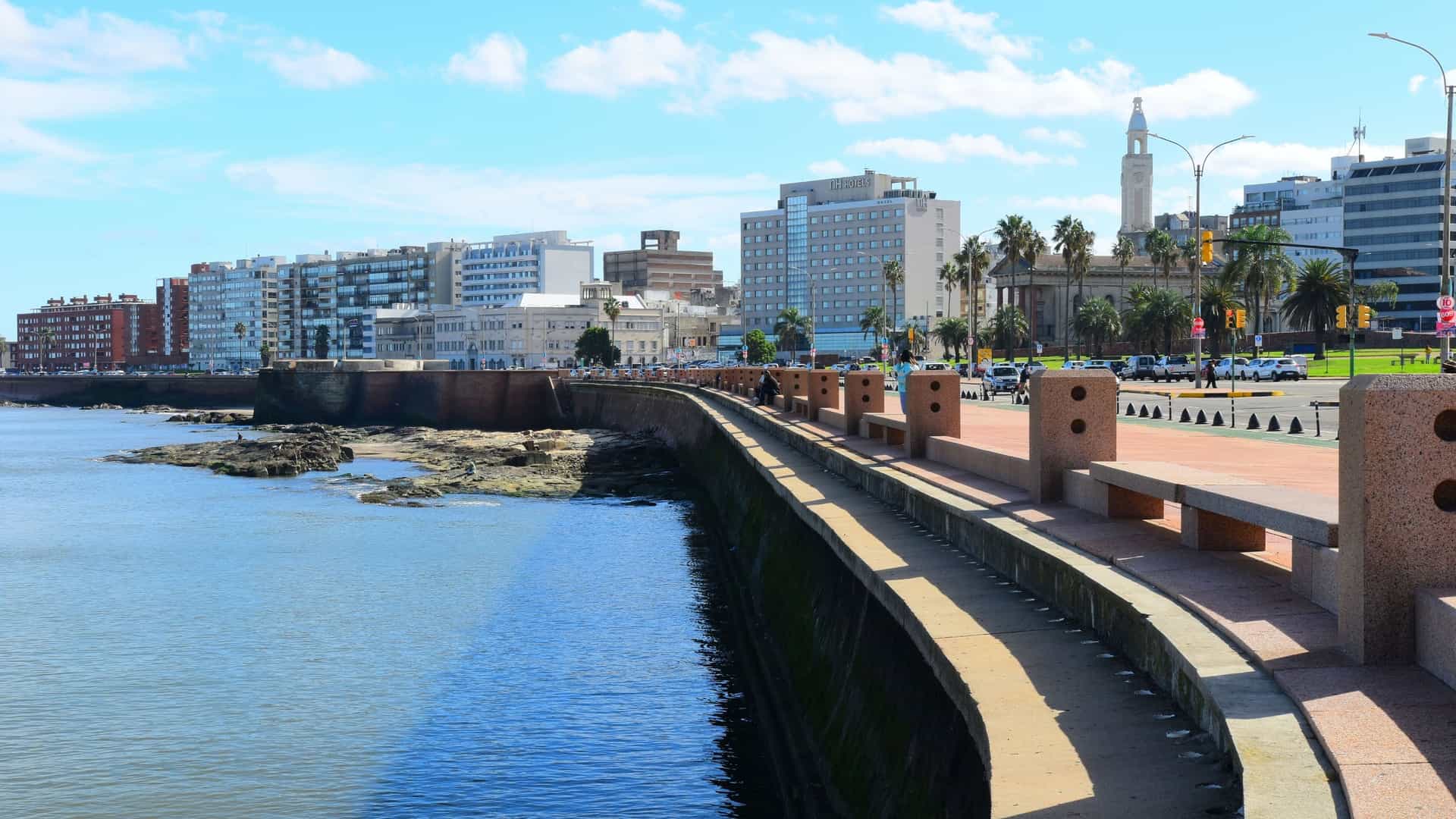 A view of Montevideo's waterfront promenade and skyline along the Rio de la Plata, with modern buildings, palm trees, and the historic Ciudad Vieja bell tower visible in the distance.
