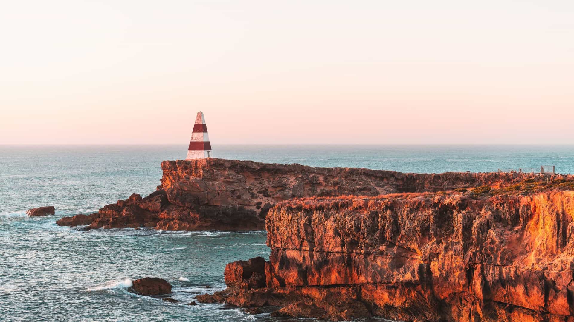 A scenic view of the Obelisk, a prominent red and white striped lighthouse, perched on a rocky cliff overlooking the sea in the coastal town of Robe, South Australia.