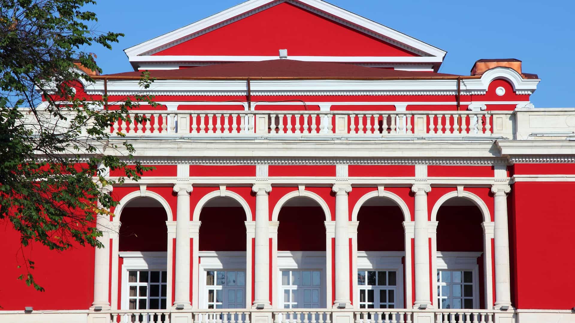 A striking close-up of the facade of the red-and-white neoclassical theater building in Rousse, Bulgaria, with large arched windows and a detailed balcony.
