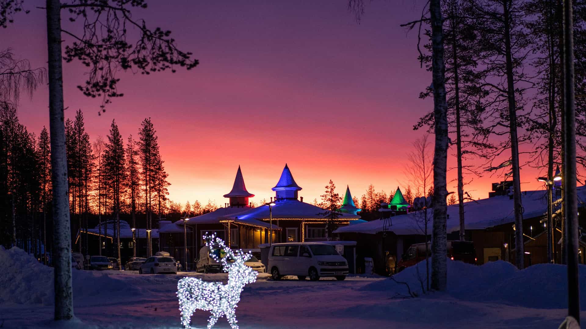 "A beautiful winter scene at the Santa Claus Village in Rovaniemi, Finland, with festive lights and a reindeer statue, set against a colorful sunset sky.  "